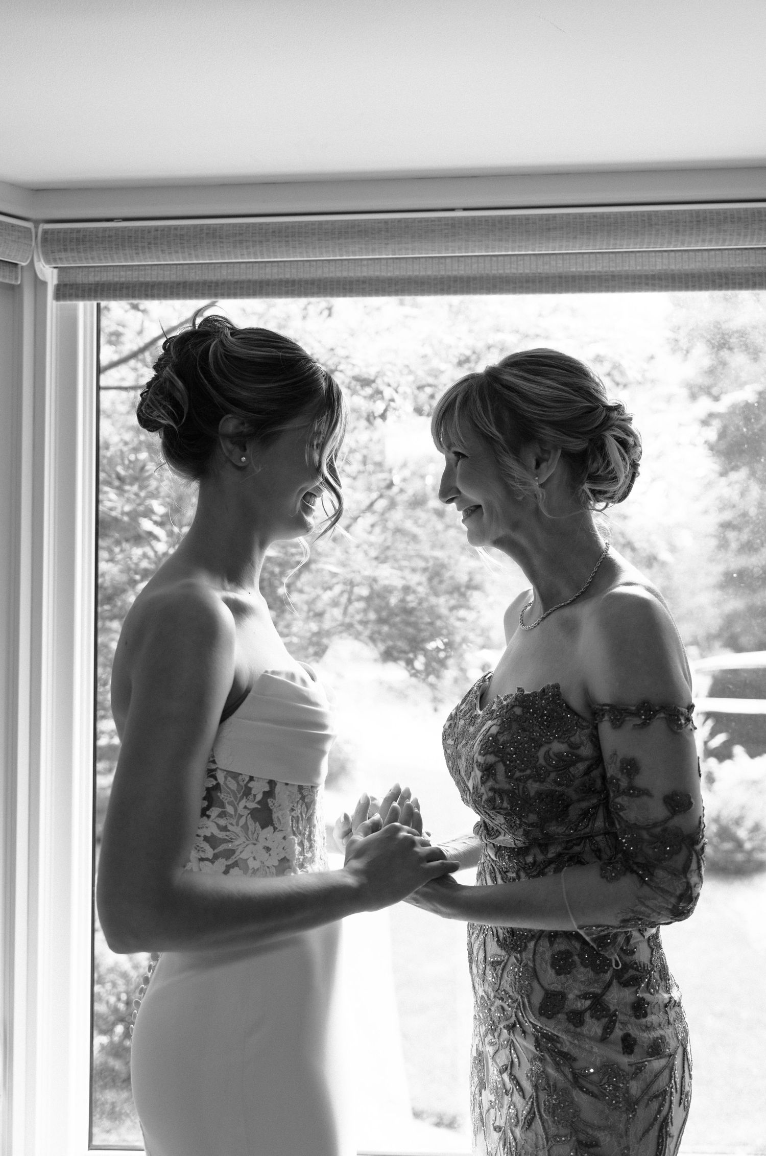 Two women in strapless dresses share an intimate moment by a window in a black and white photograph.