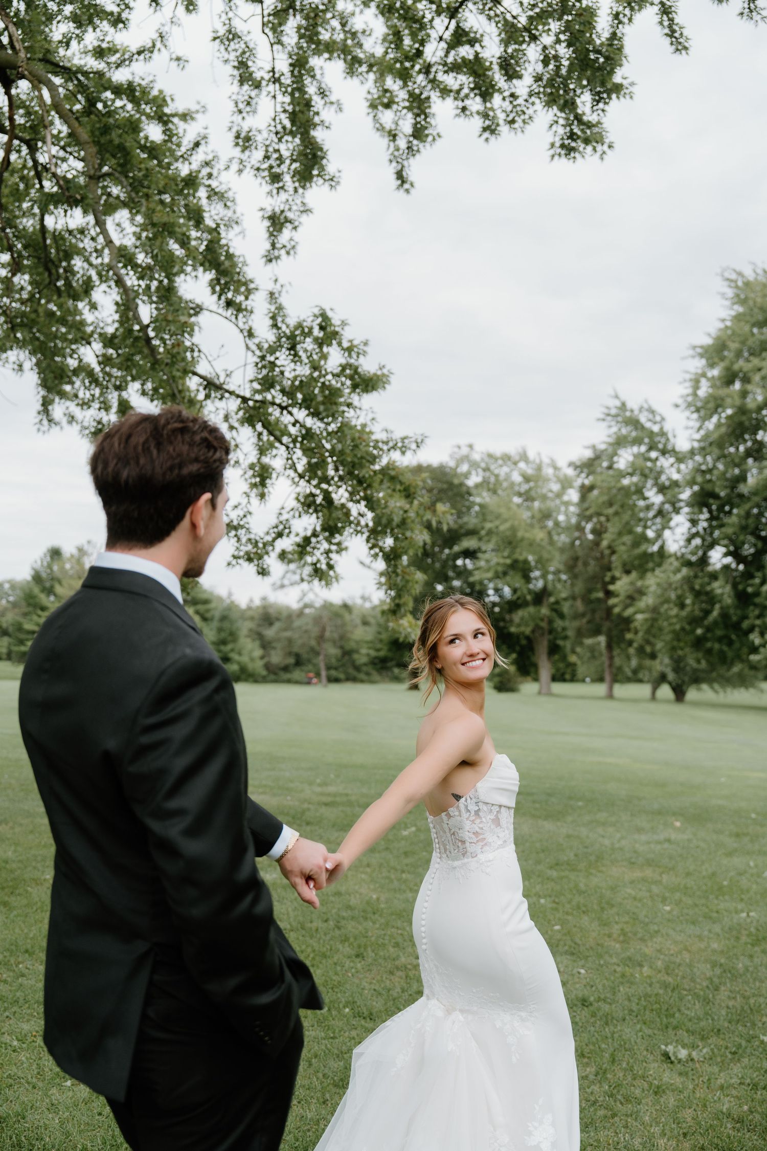 A couple in wedding attire dances and twirls together on a grassy lawn surrounded by trees.