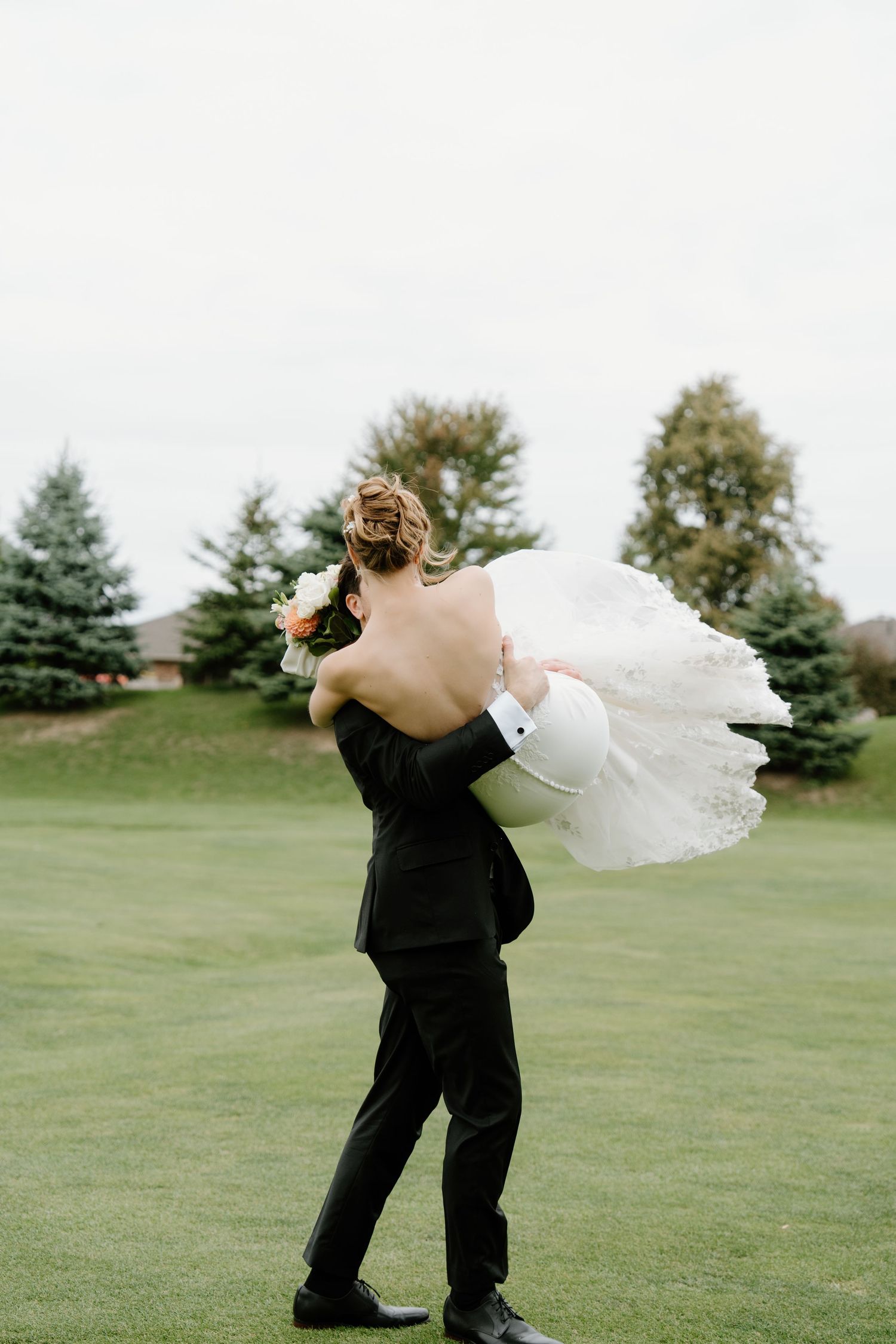 A couple shares a romantic embrace on a lush green golf course with their white wedding dress flowing in the wind.