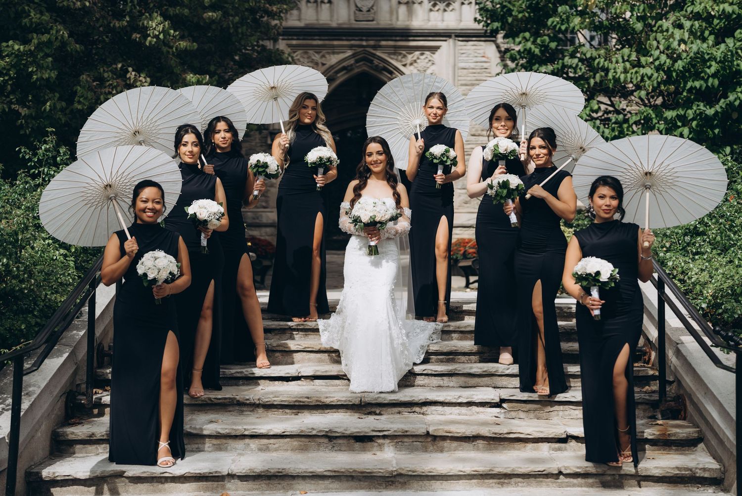 Bridal party with white parasols and dark gowns gathered on historic stone staircase for formal photo.