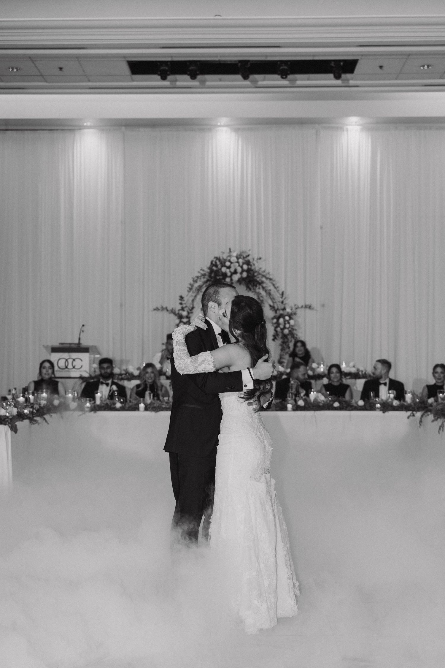 A couple shares their first dance as newlyweds in a white-walled reception hall with guests watching from their tables.