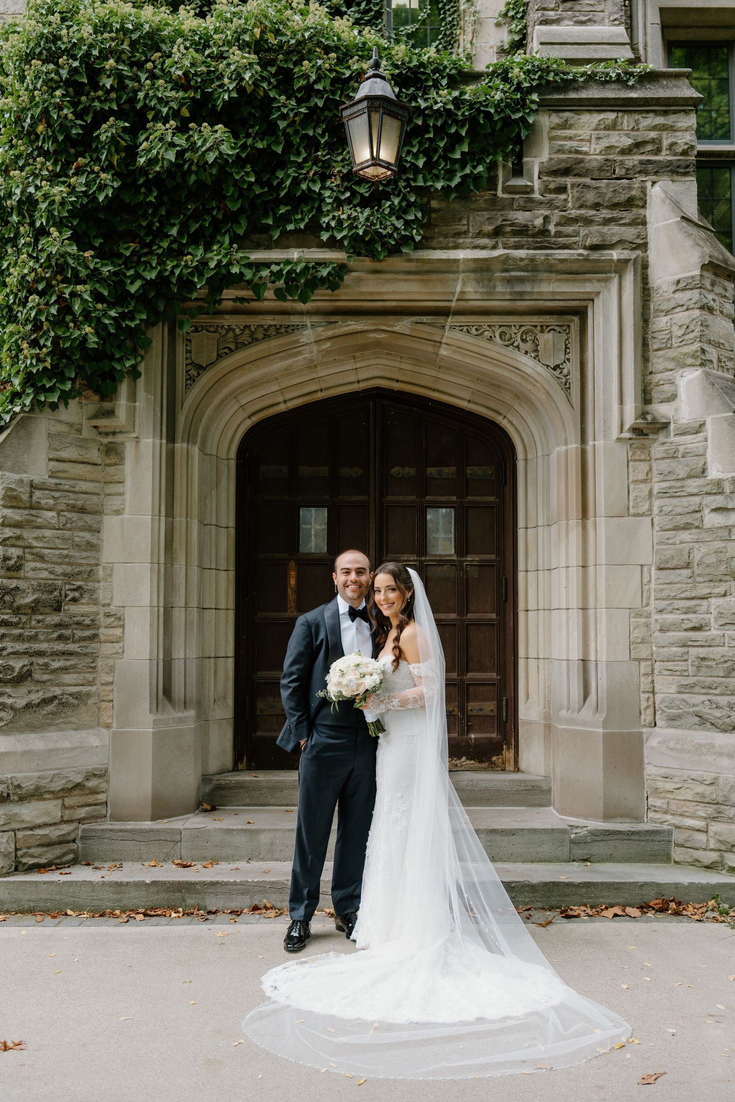 A couple poses in formal wedding attire under a Gothic stone archway with ivy growing on the building.