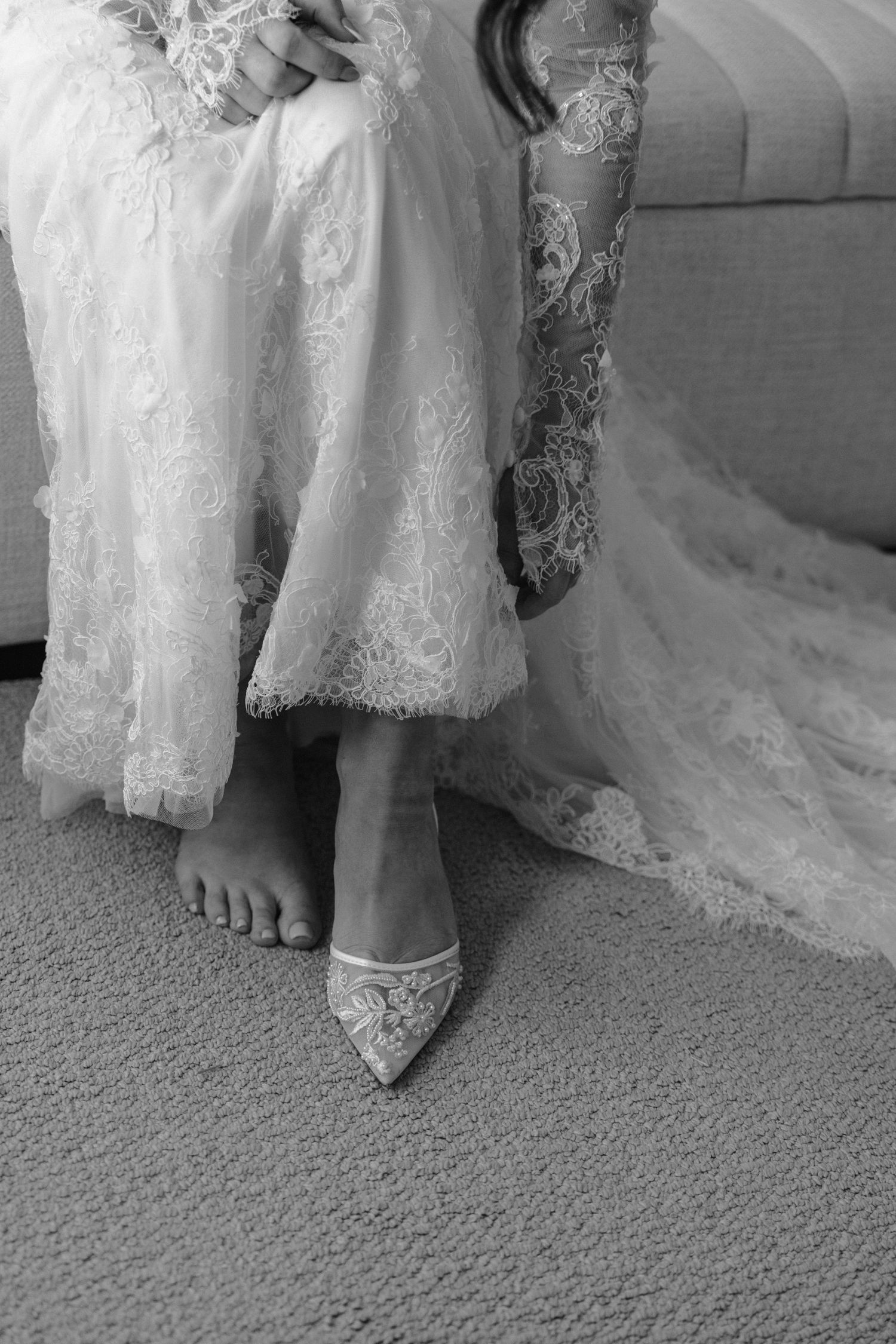 Close-up view of bare feet peeking out from under delicate white lace wedding dresses on a carpeted floor.