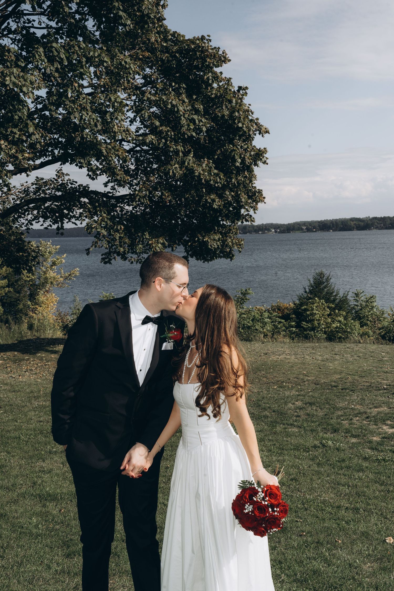 A couple in wedding attire share a romantic moment on a grassy hillside overlooking a serene lake.