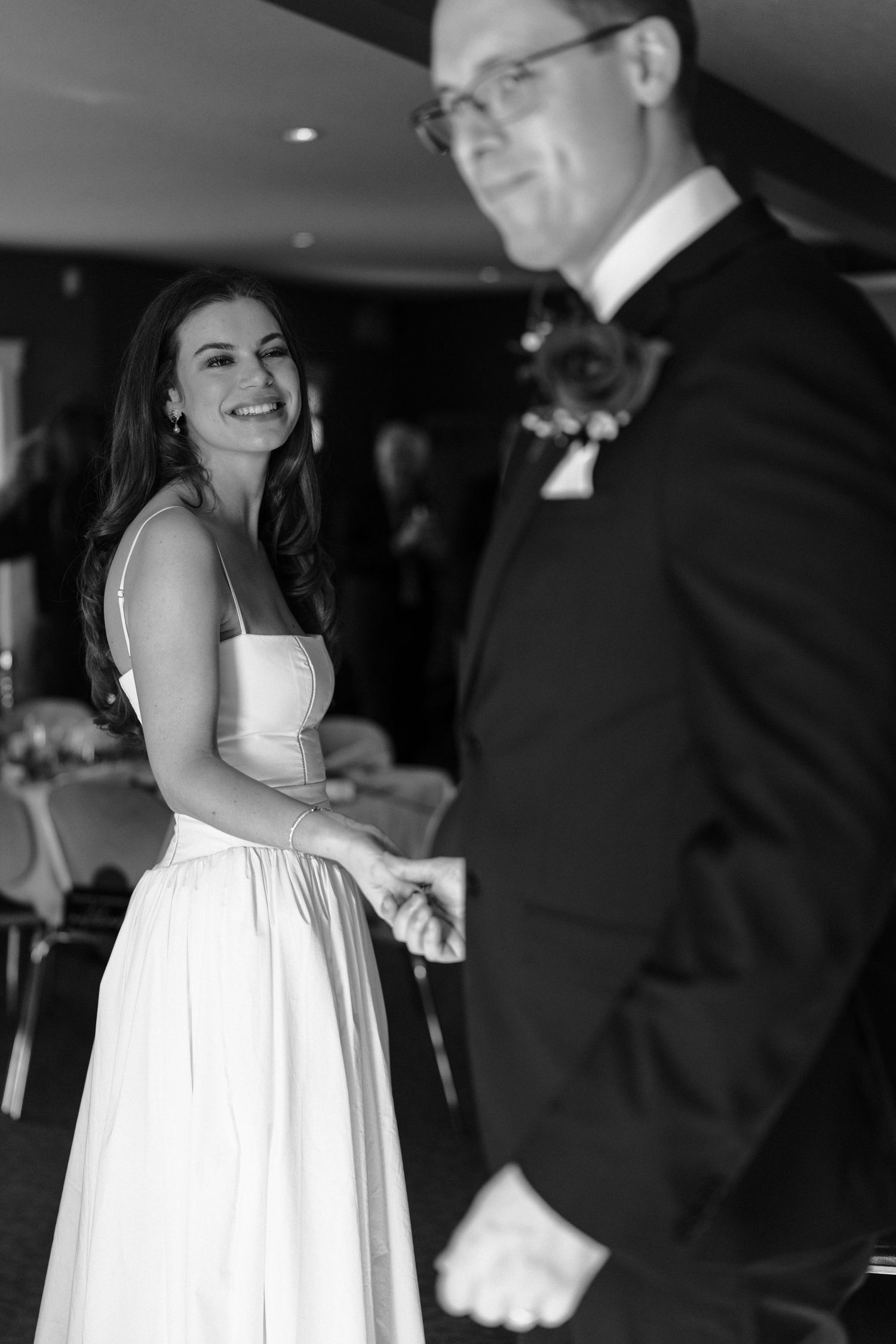 A bride in a white dress and groom in a tuxedo share a candid moment in black and white at their wedding reception.