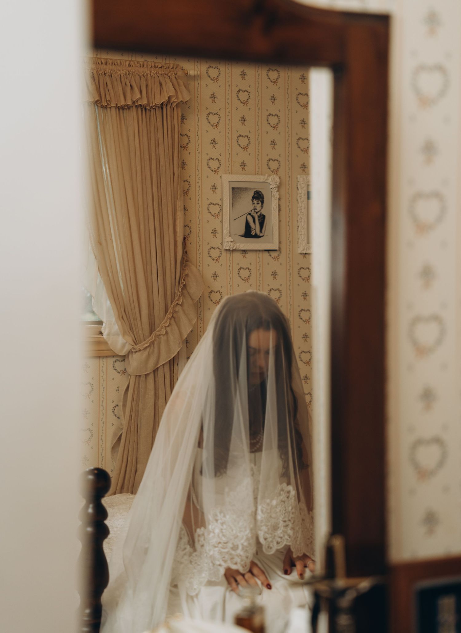 A bride with a lace veil gazes into a mirror in a room with vintage wallpaper and curtains.
