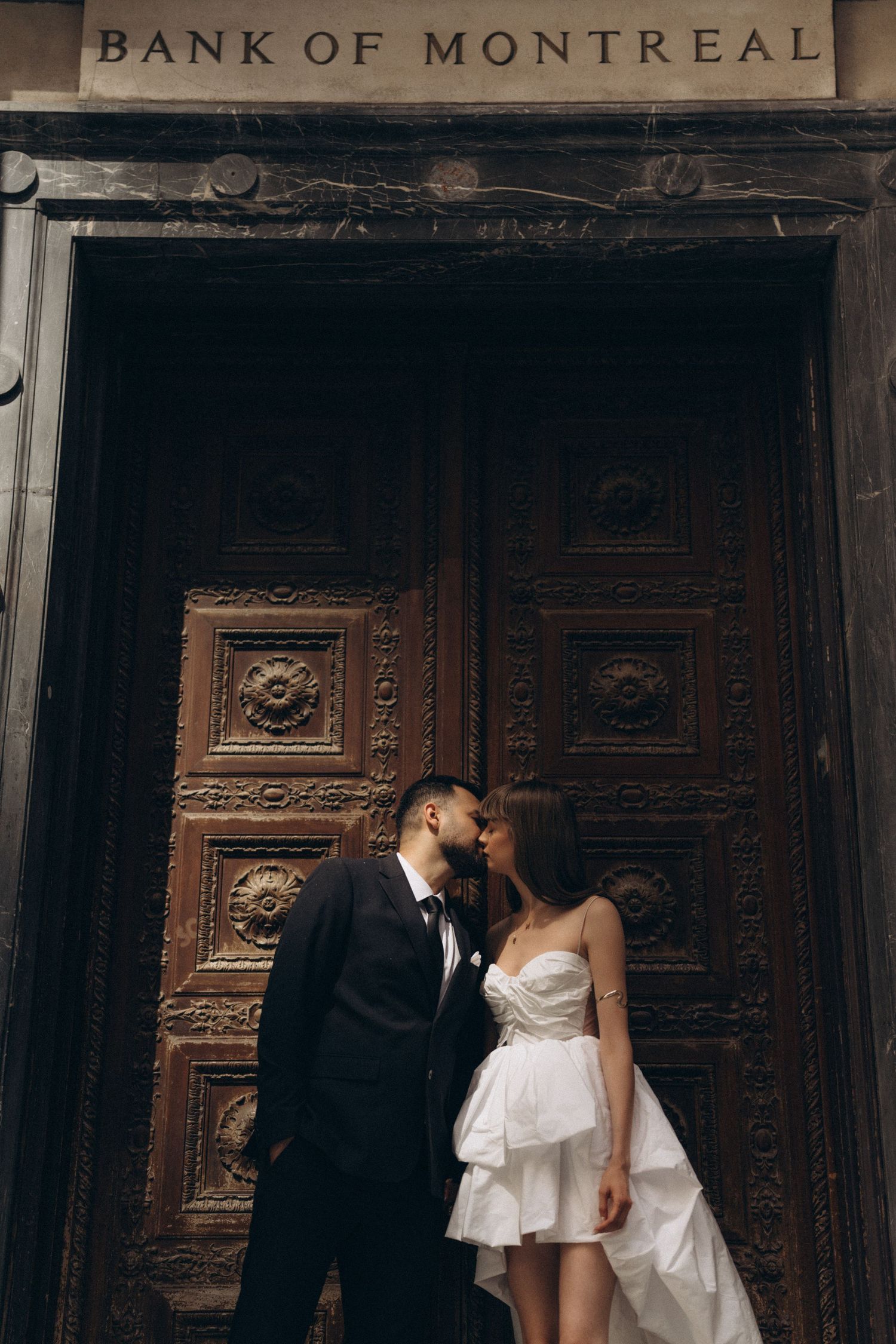 A romantic couple in wedding attire share an intimate moment in front of ornate wooden doors at Bank of Montreal.