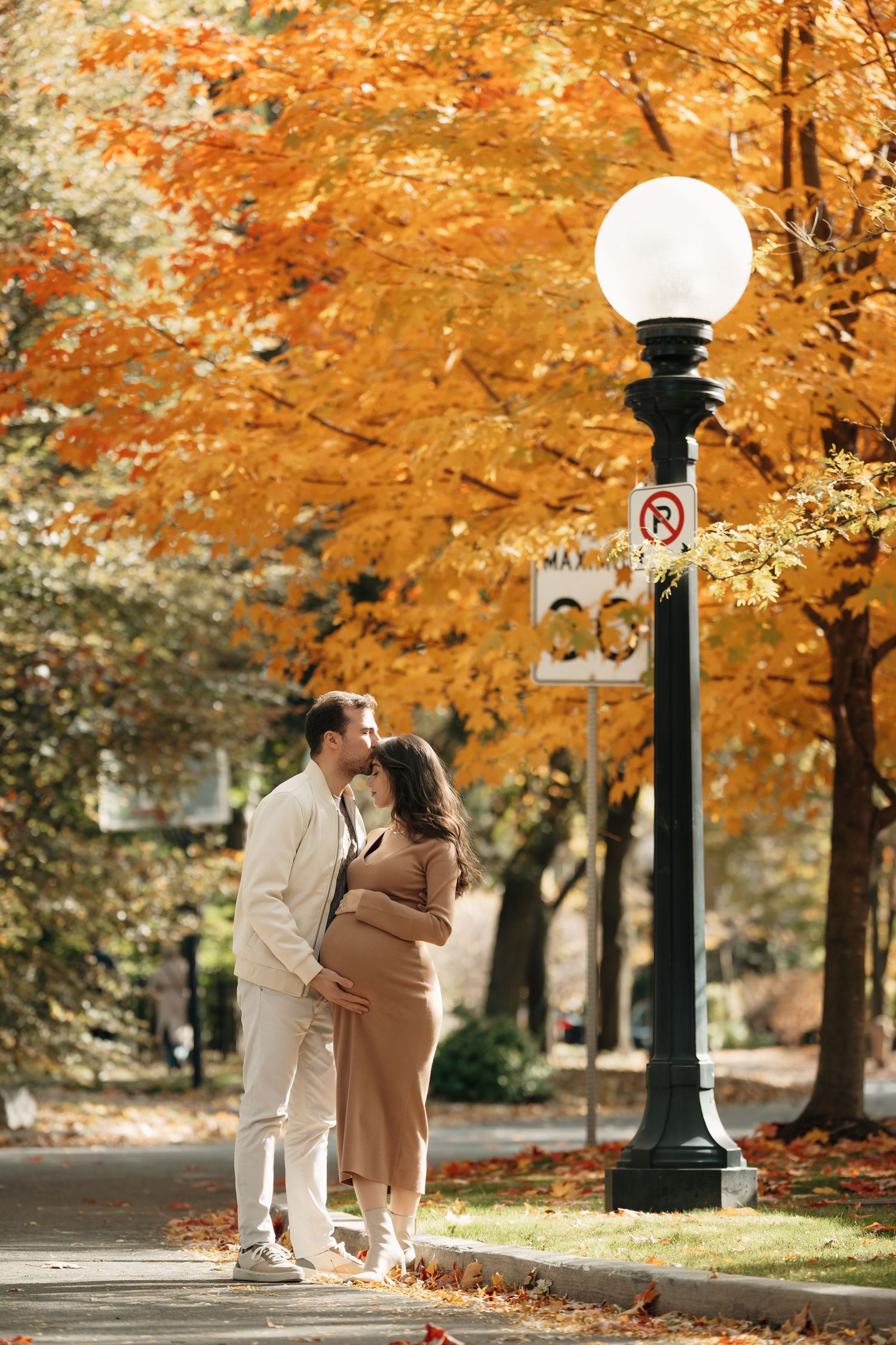 A couple shares a romantic moment surrounded by golden fall foliage and lamp posts in an autumn park setting.