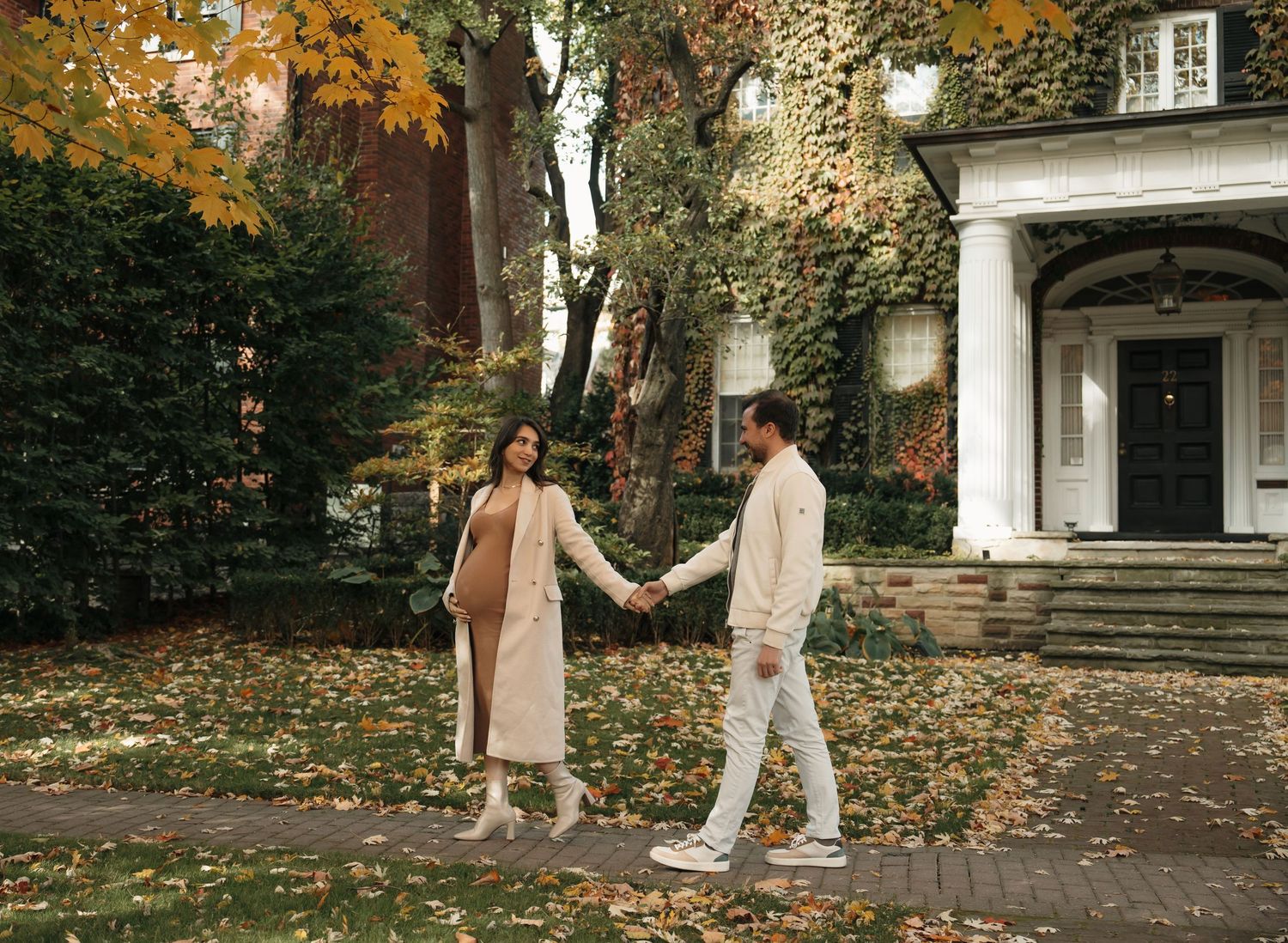People walking along a tree-lined path covered in fallen autumn leaves with historic architecture in the background.