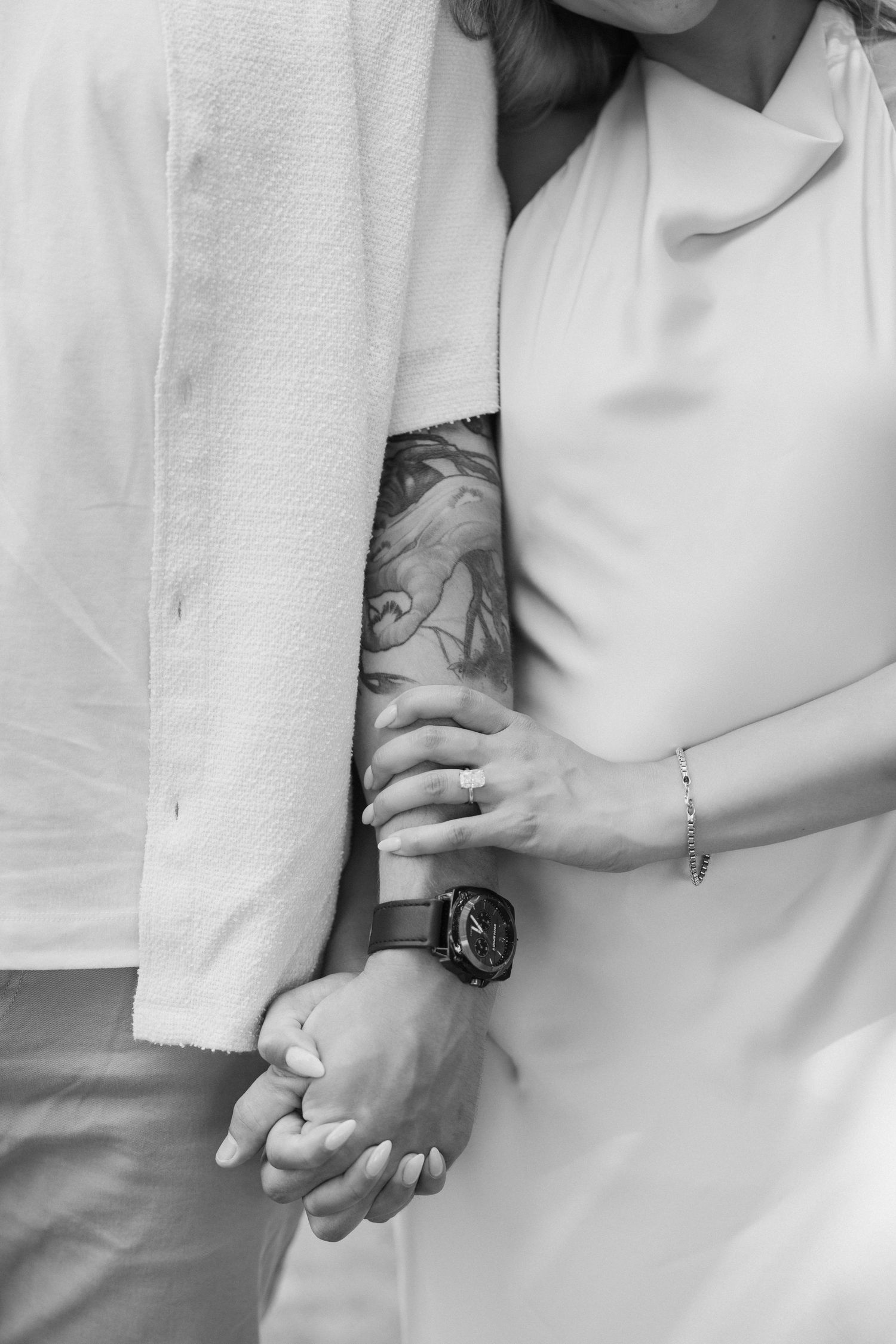 A black and white close-up of hands holding each other, featuring tattoos and a wristwatch.