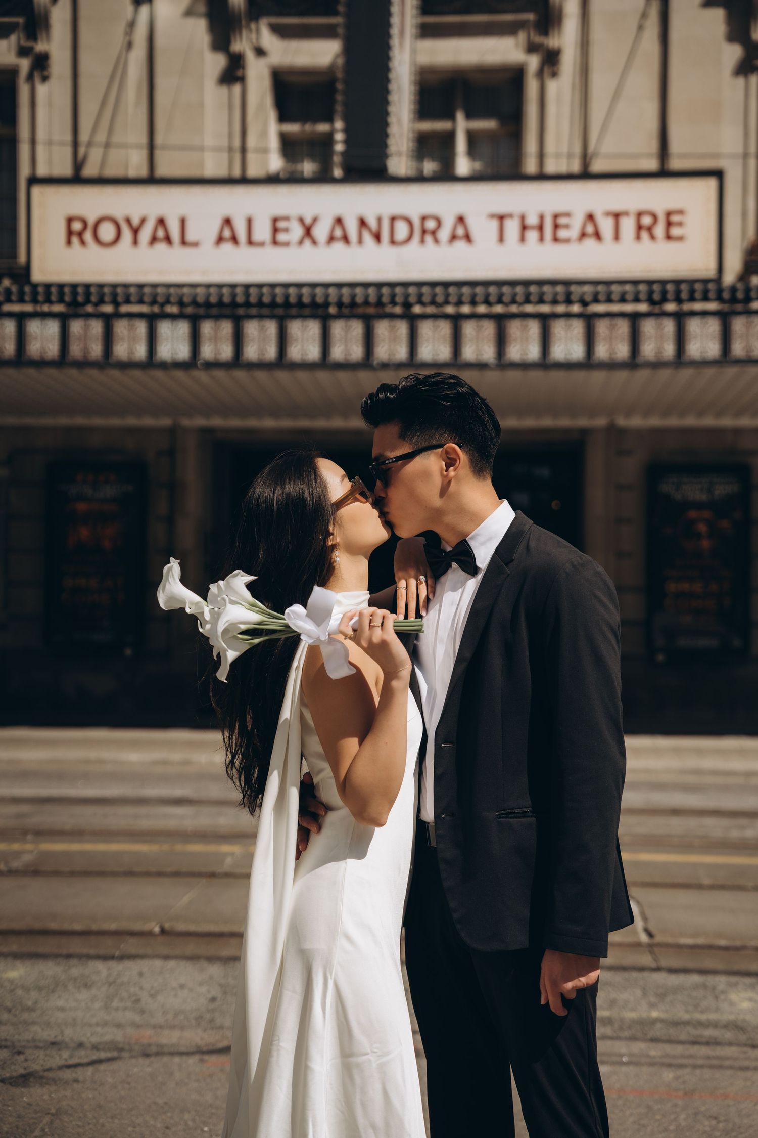 A wedding couple shares an intimate moment outside the Royal Alexandra Theatre on an overcast day.