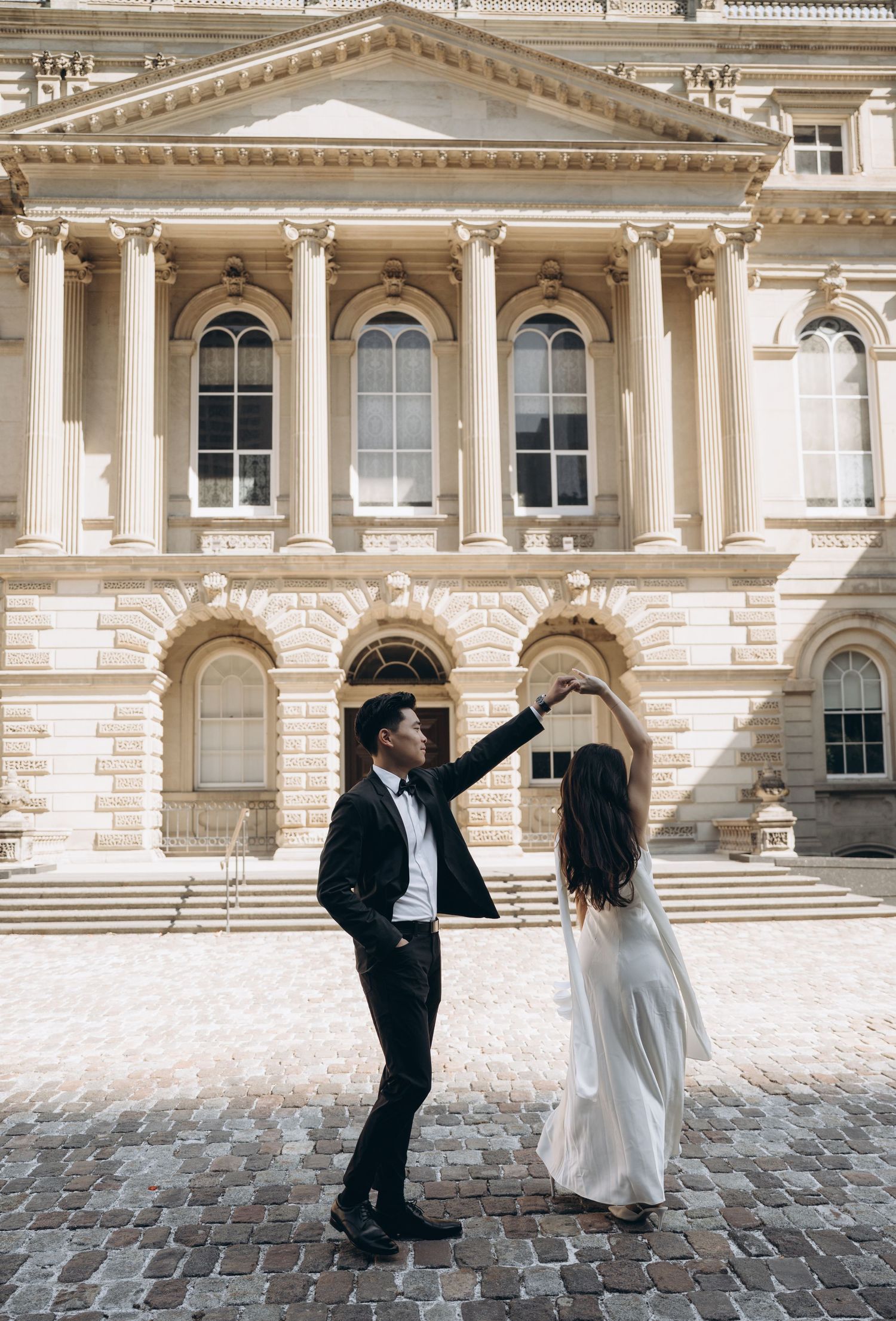 A couple dancing on cobblestones in front of a grand neoclassical building with white columns and arched windows.