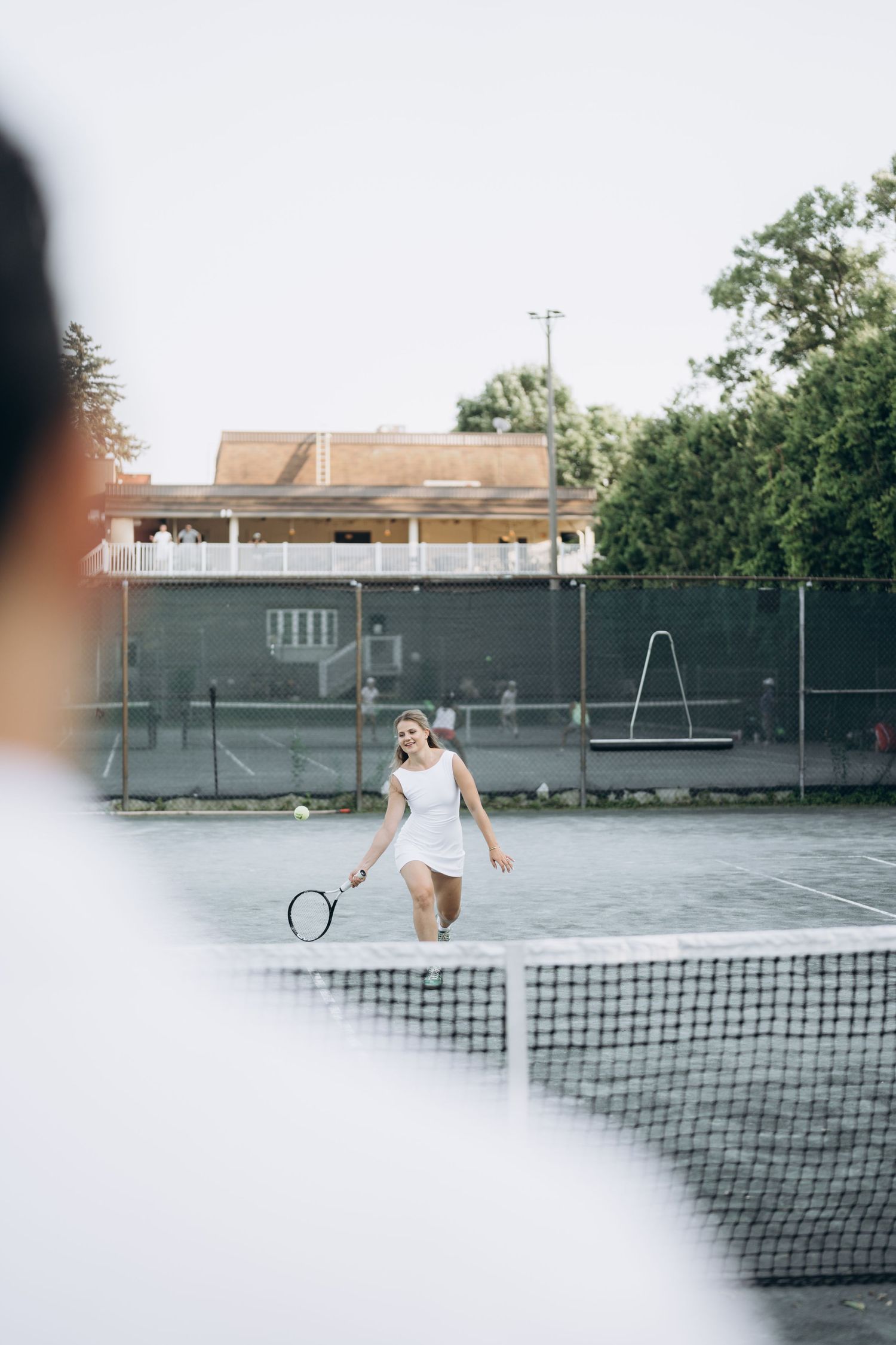 Players engage in a tennis match on an outdoor court with green surroundings and net visible.