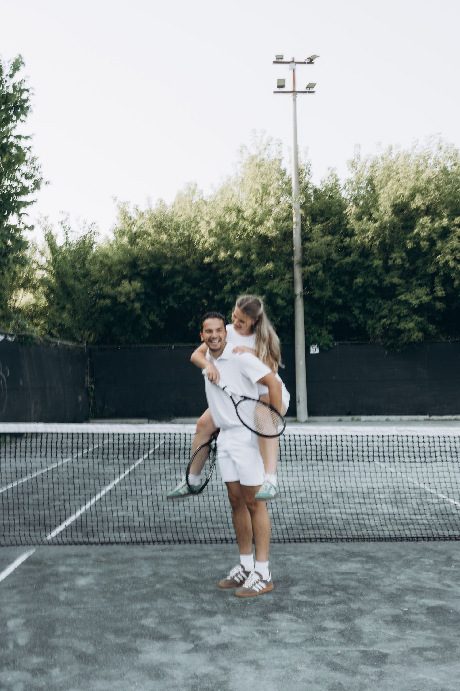 Athletic couple embracing on tennis court with rackets and net, wearing white sportswear on sunny day.
