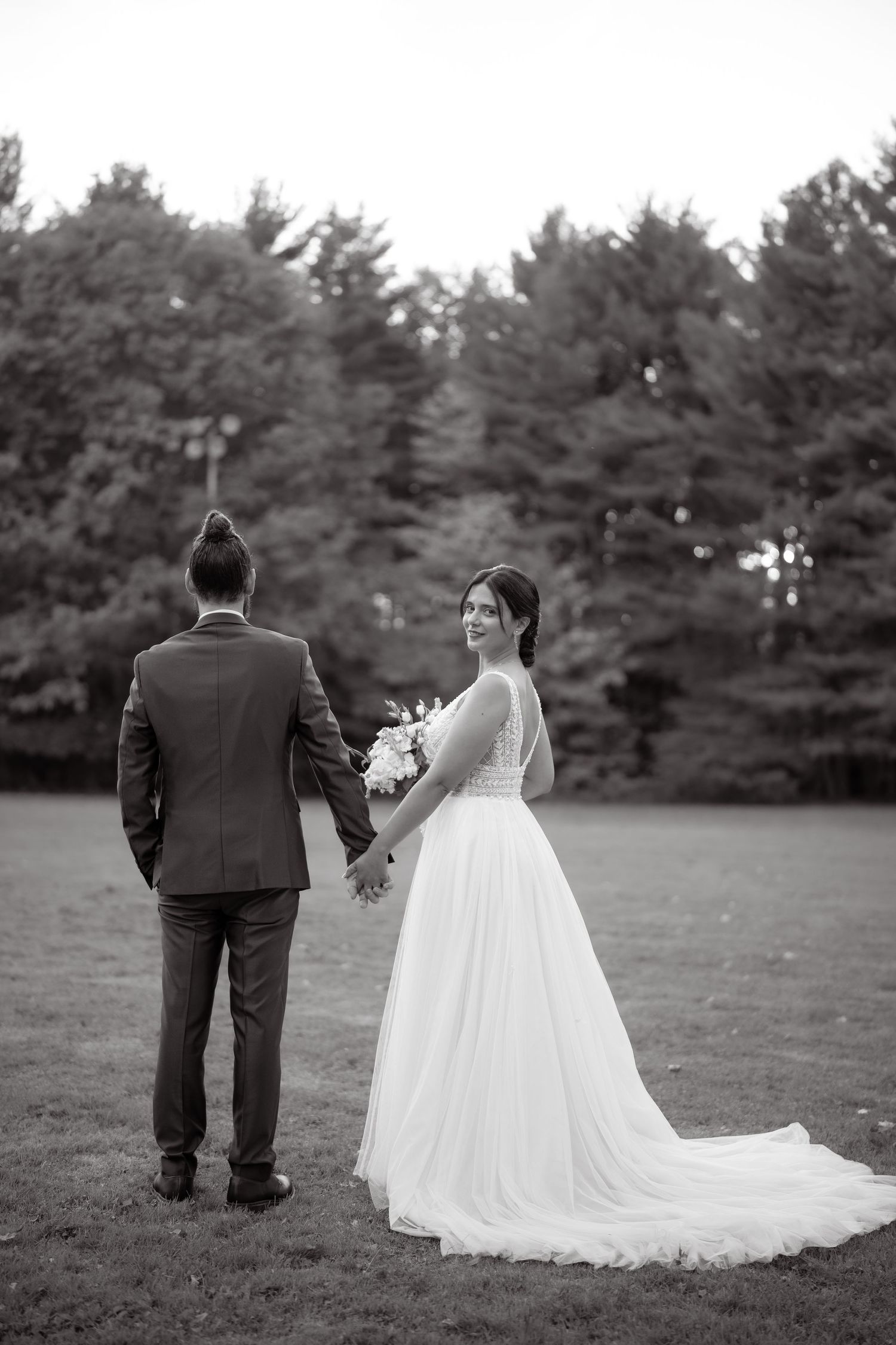 Black and white wedding portrait of couple standing on lawn with pine trees in background.
