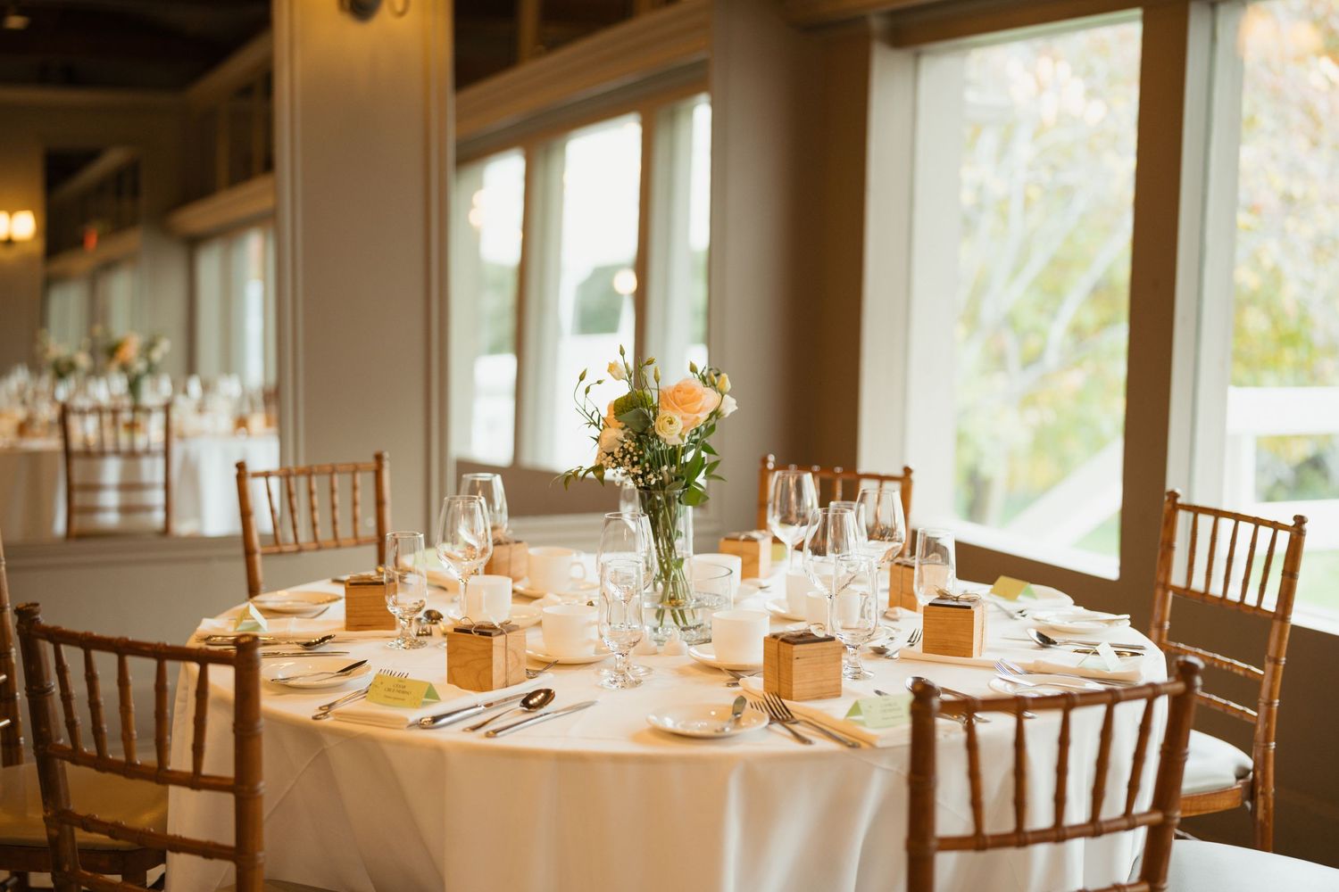 Bright reception venue with round tables featuring elegant place settings and warm window lighting.