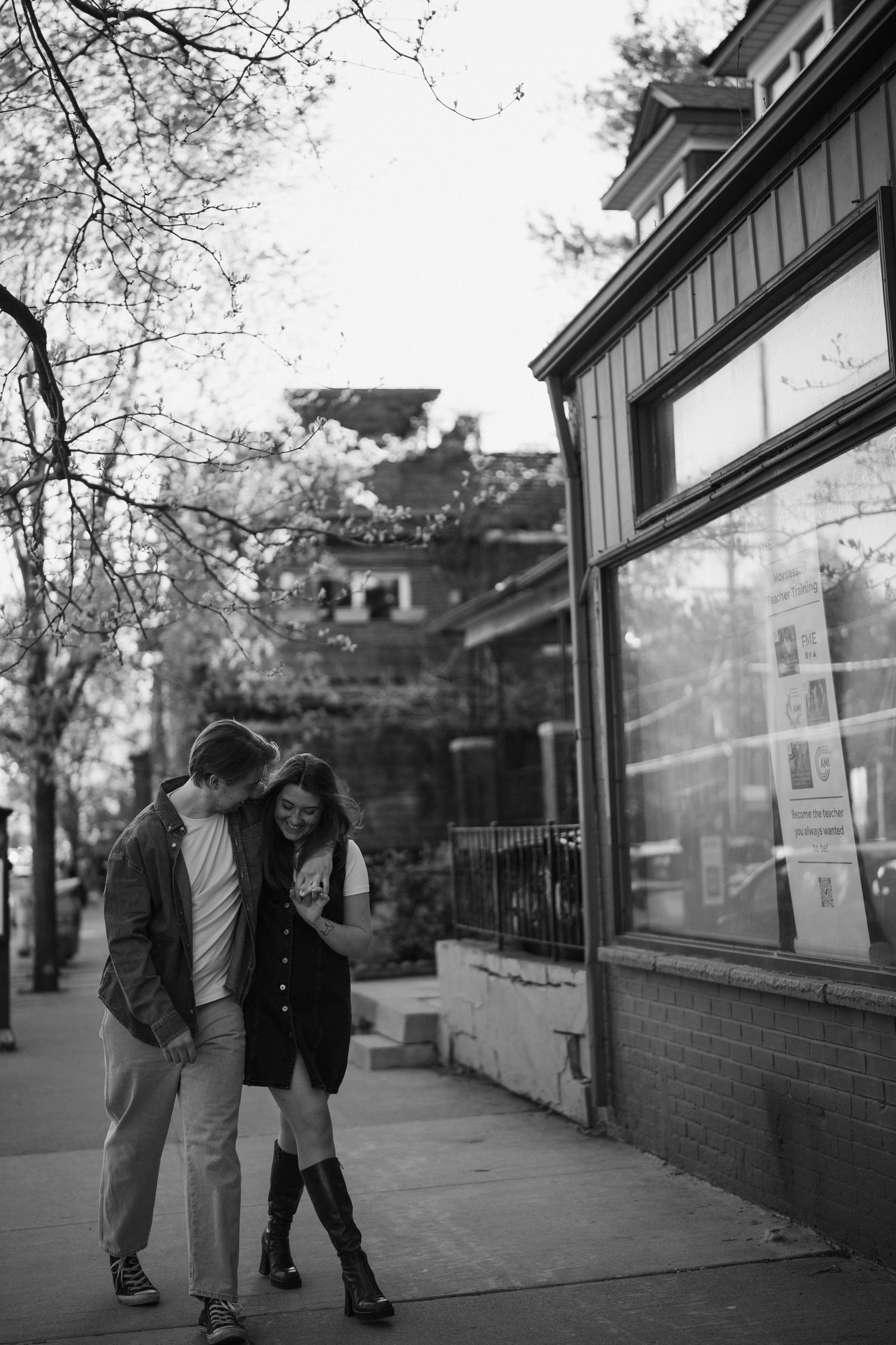 A couple embraces on a city sidewalk next to a storefront in black and white photography.