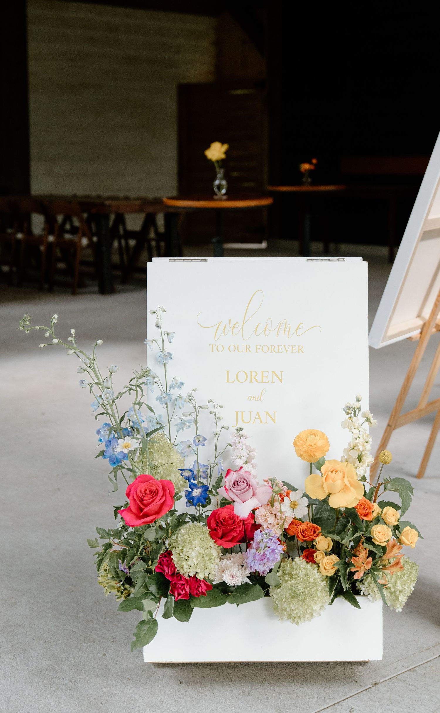 Elegant white wedding welcome sign with gold lettering surrounded by colorful floral arrangement of roses and hydrangeas.