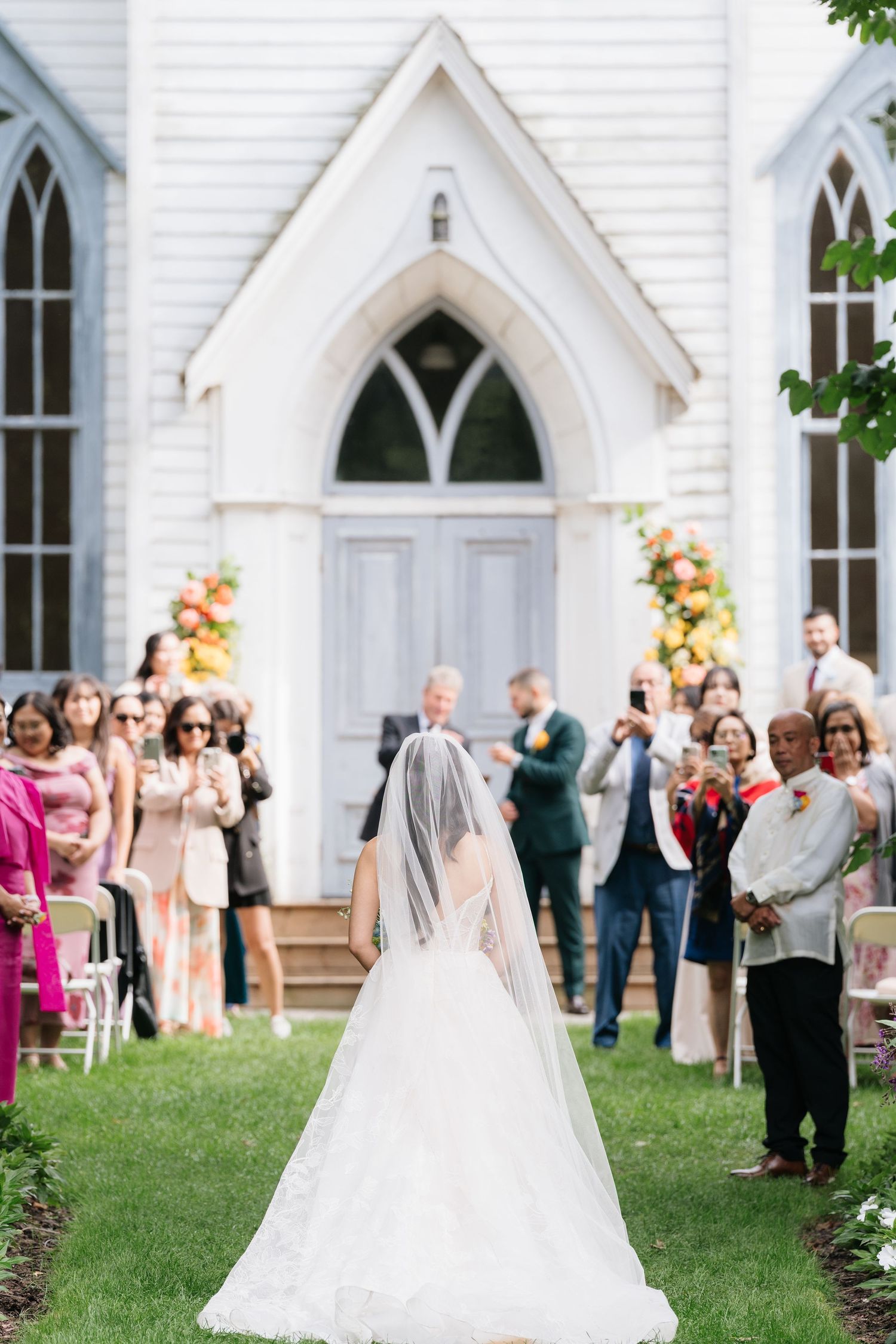 Wedding guests gather outside a charming white Gothic-style church with arched windows and doors.