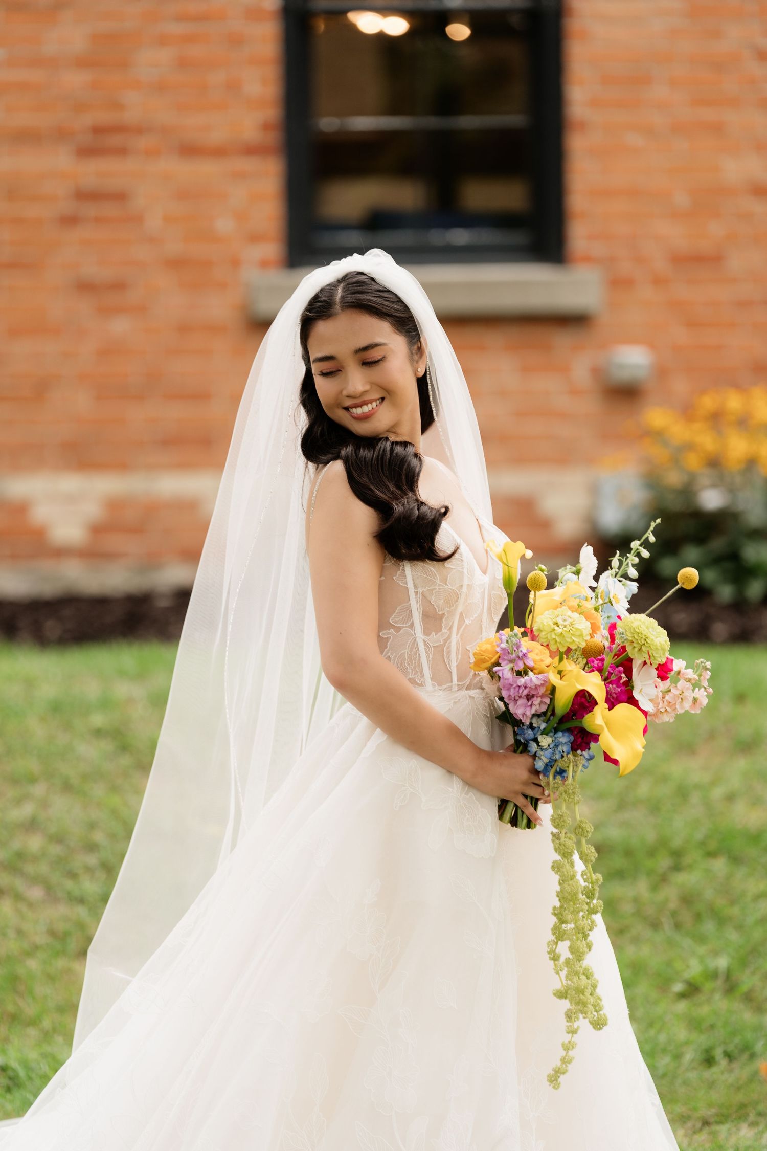 A bride in a white wedding dress holds a vibrant bouquet with yellow and pink flowers against a brick wall.