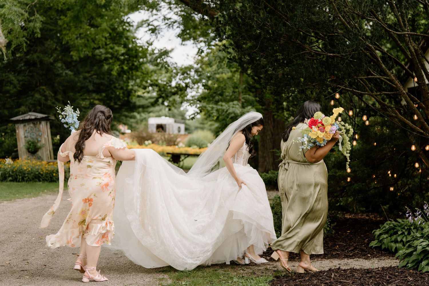 Wedding party members help carry a flowing white wedding dress across a garden path.