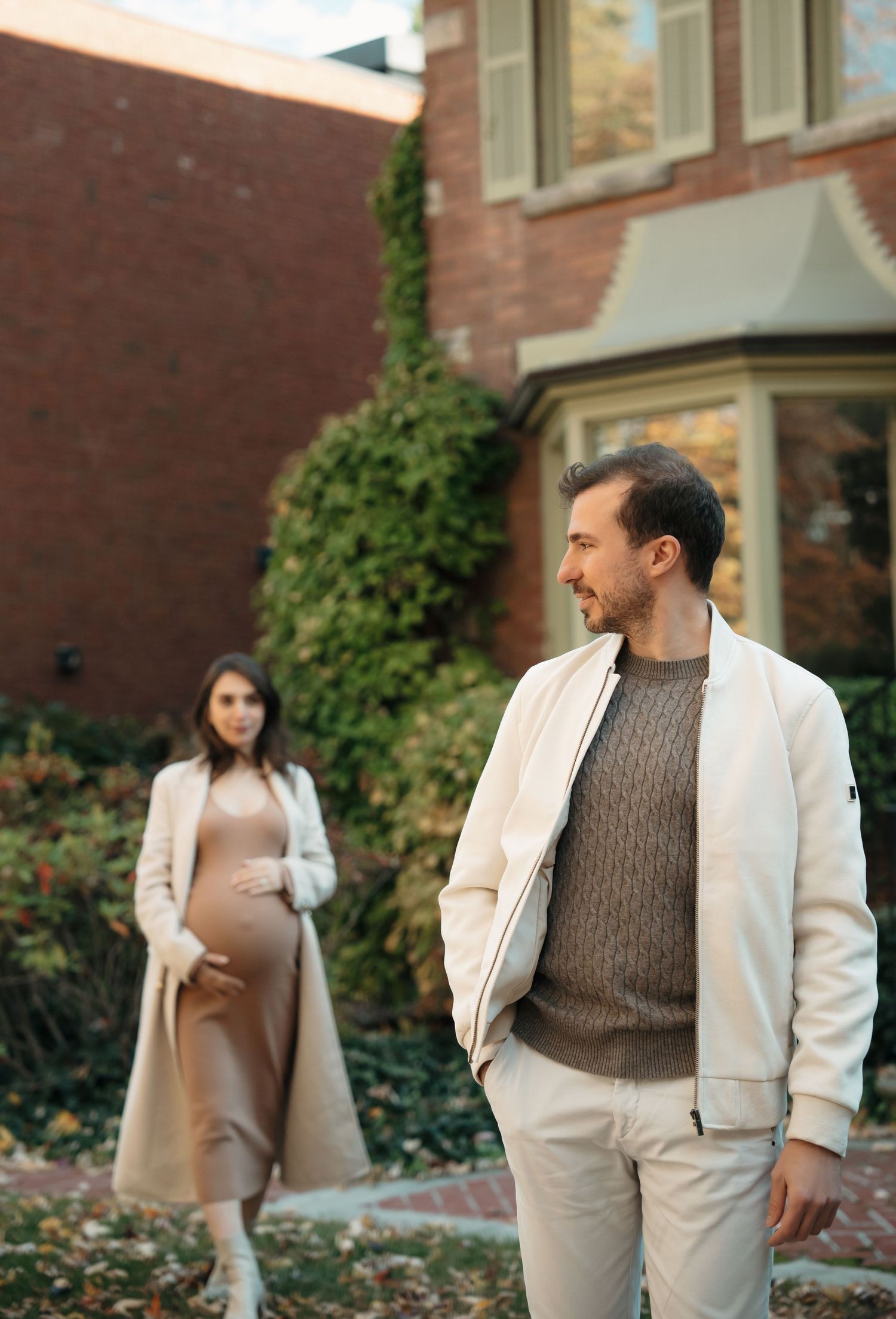 Couple in coordinated cream and brown outfits pose together against brick building with climbing vines.