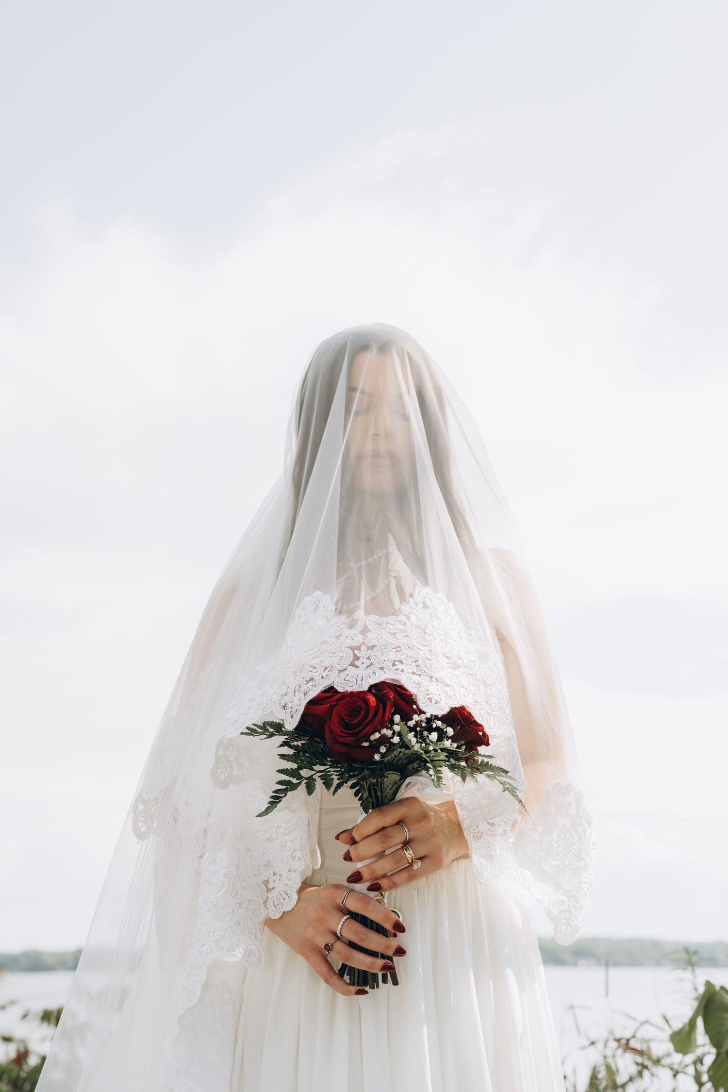 Bride covered in delicate veil clutches red rose bouquet while posing outdoors against cloudy backdrop.