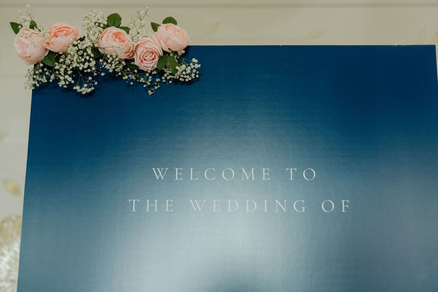 A wedding welcome sign with pink roses and floral decorations against a blue gradient background.
