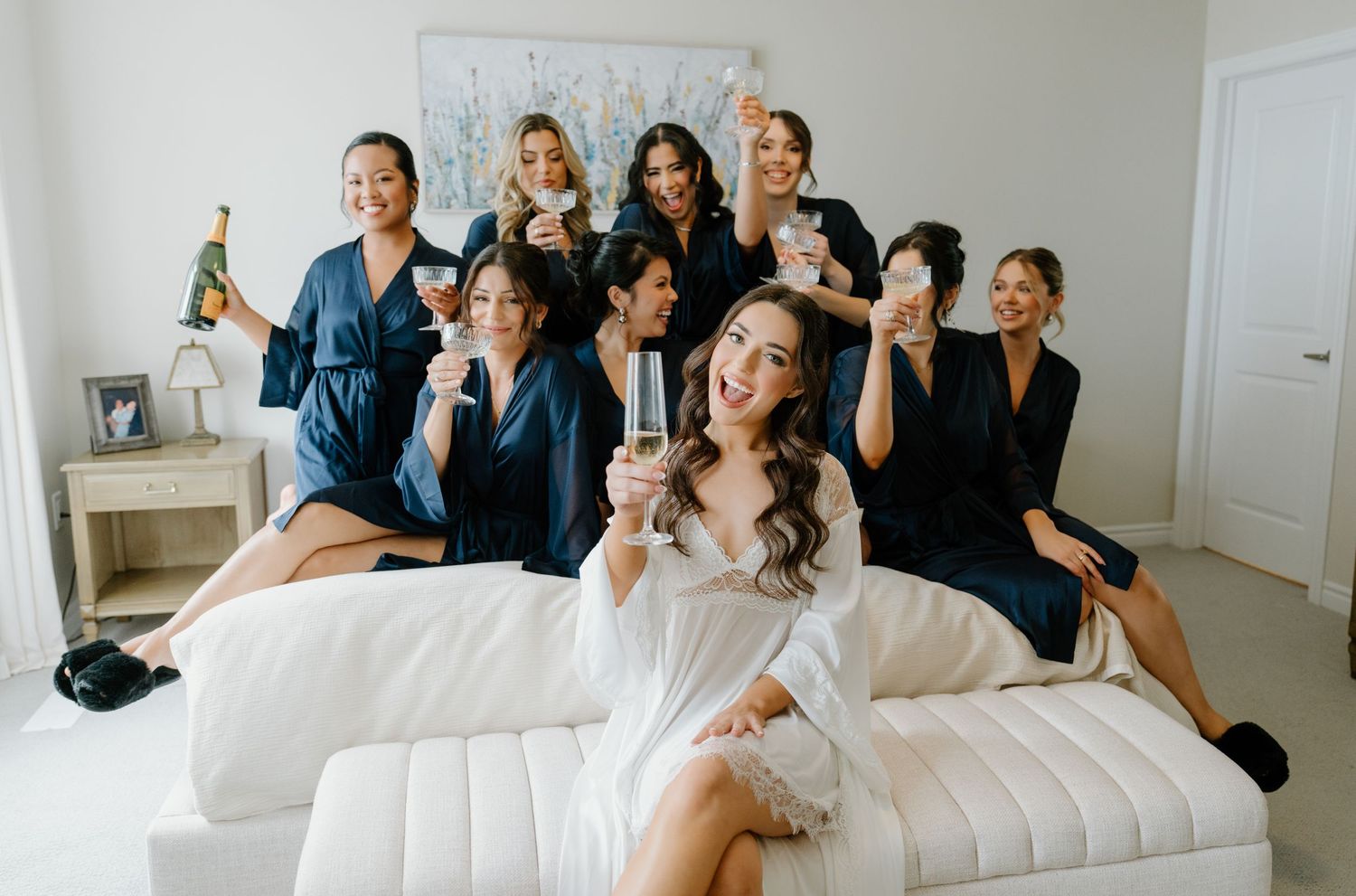 Wedding party wearing navy blue dresses lounges together on a white sofa during a celebratory moment.