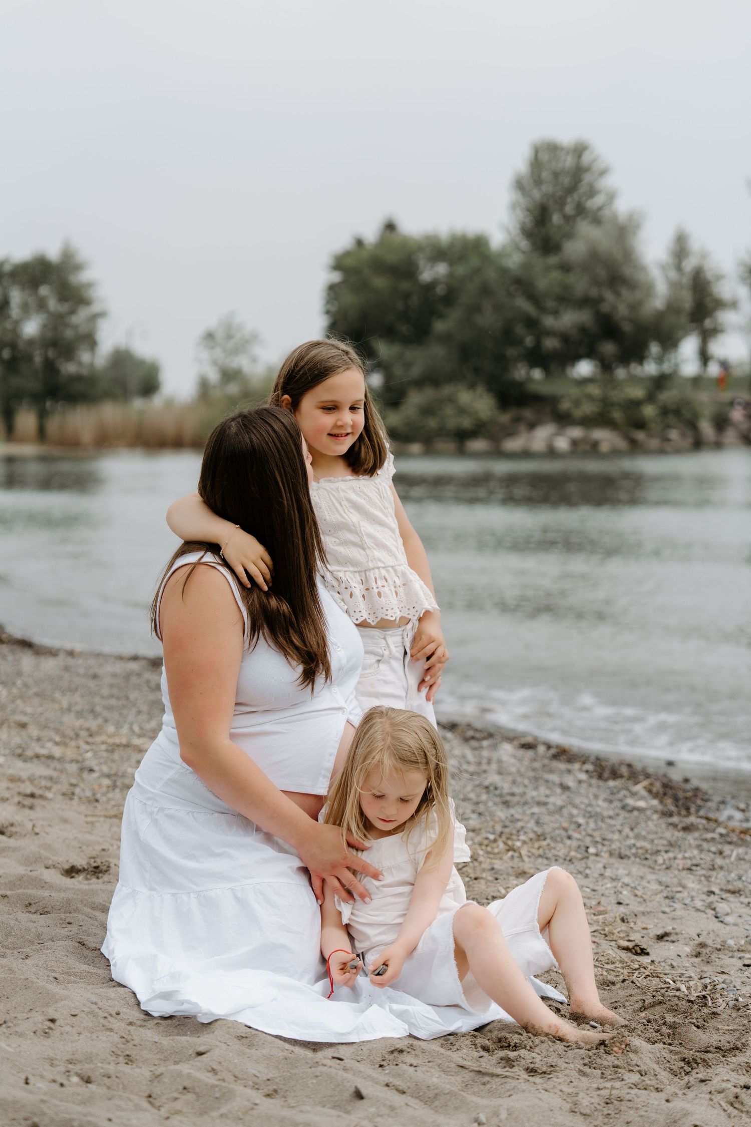 Family wearing white dresses sits together on a beach shore near the water on an overcast day.