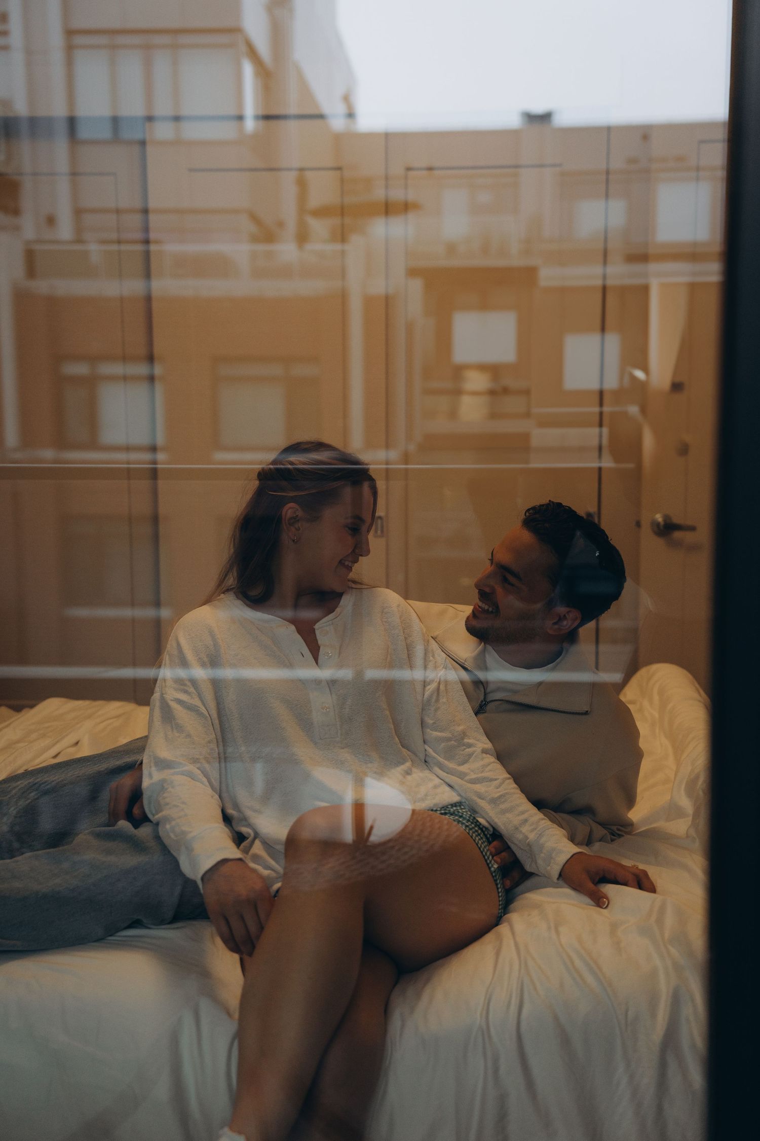 Two people having an intimate conversation while lounging on white bedding in natural afternoon light.