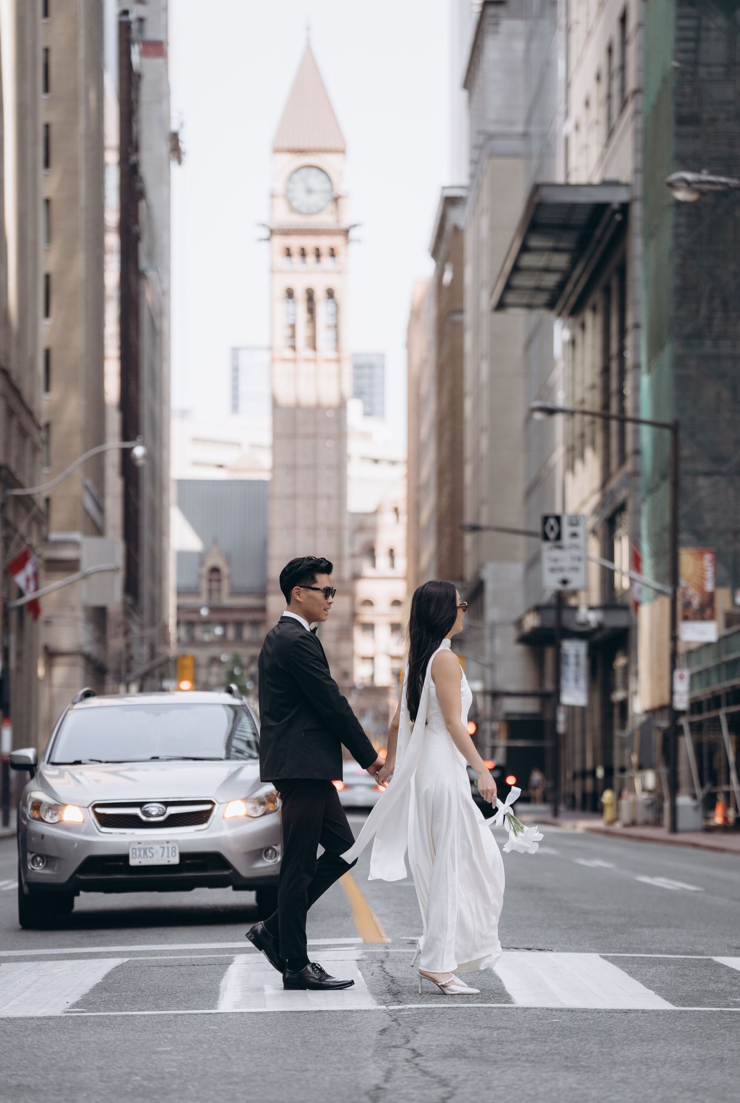 A stylish couple crosses a downtown street with a historic clock tower visible in the background.