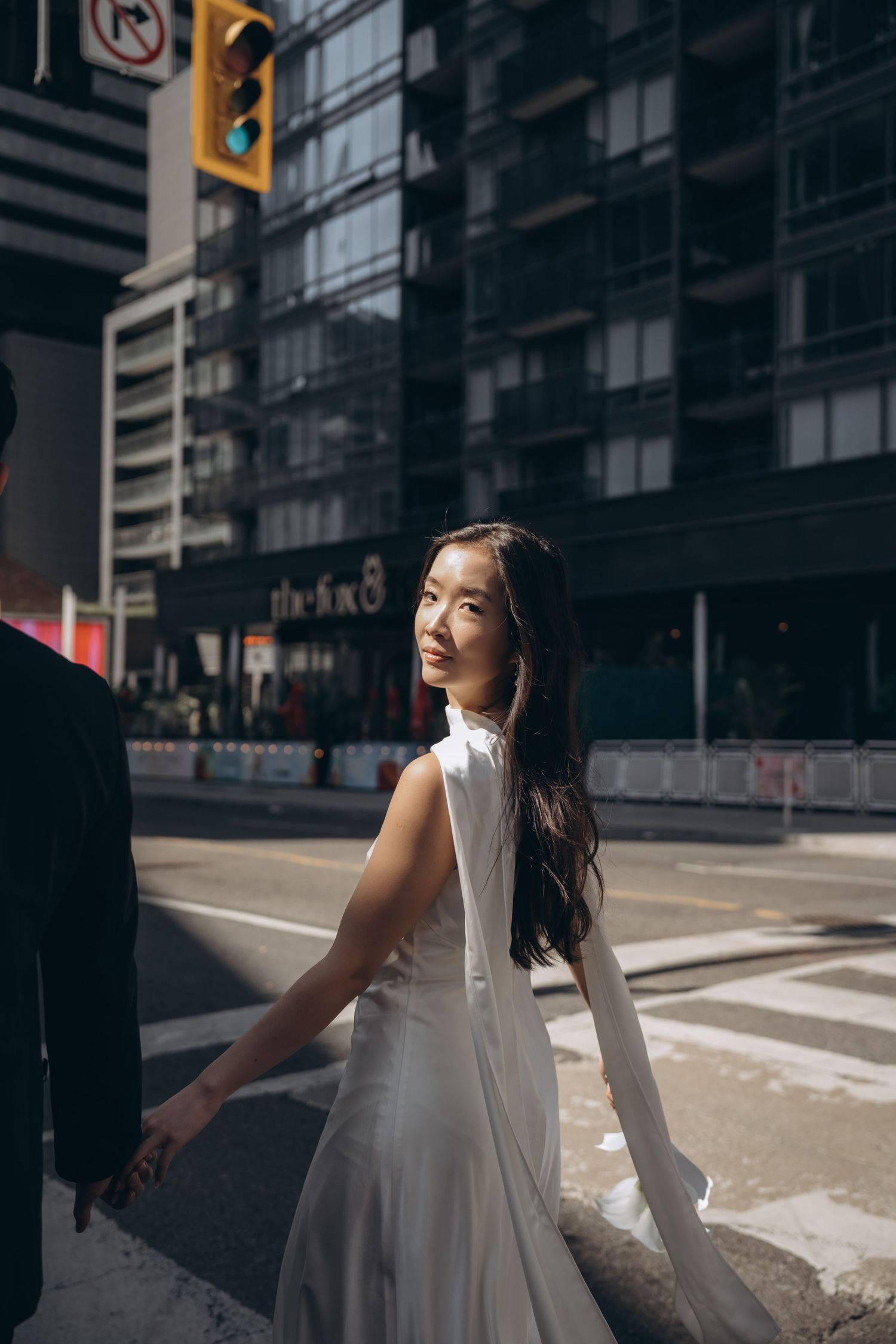 Person in an elegant white sleeveless dress poses on urban street corner with city buildings in background.