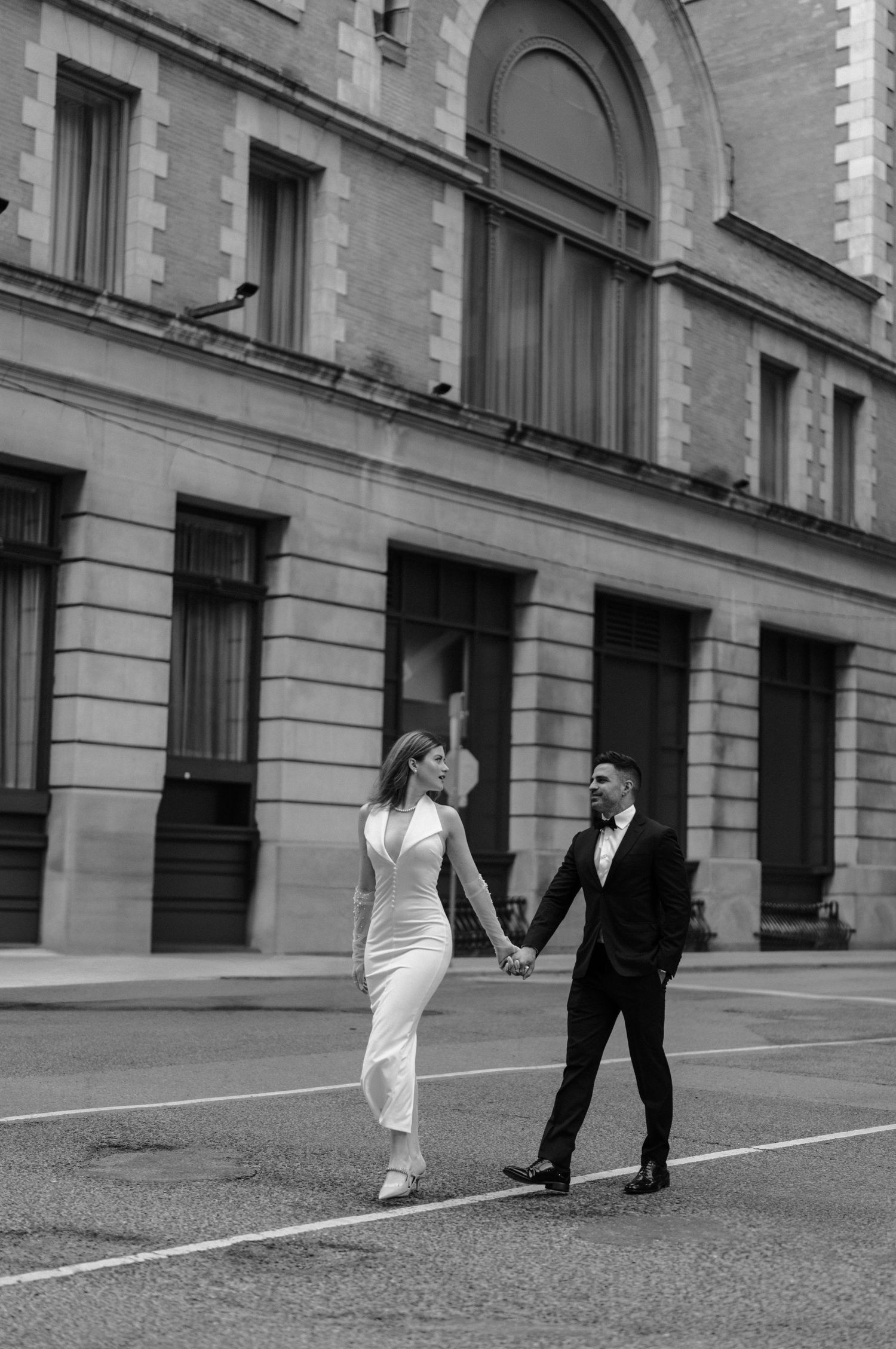 A romantic black and white photo of a couple in formal attire walking hand in hand on a city street.