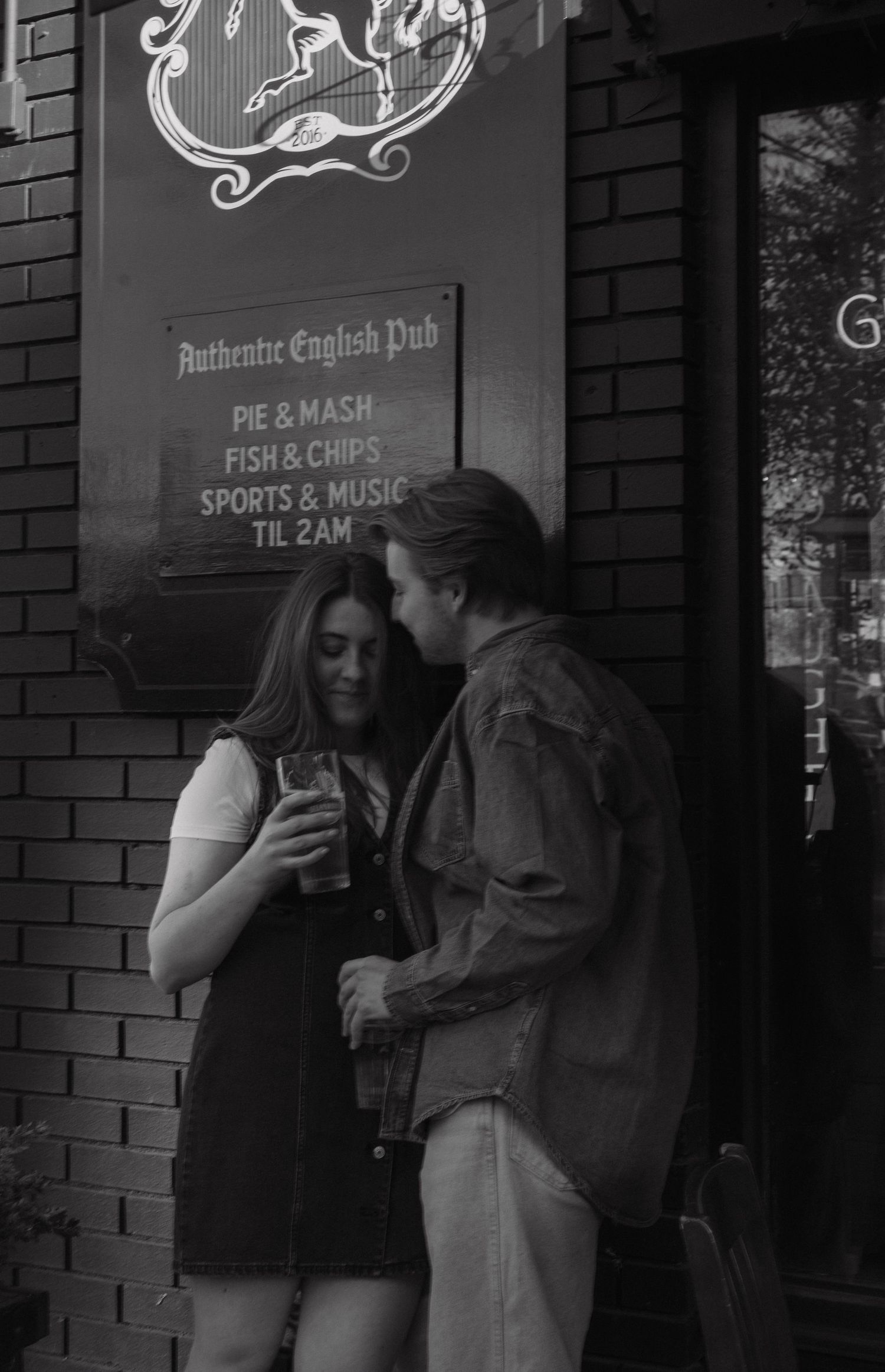 Two people chat outside an authentic English pub advertising pie and mash, fish and chips, and sports and music.