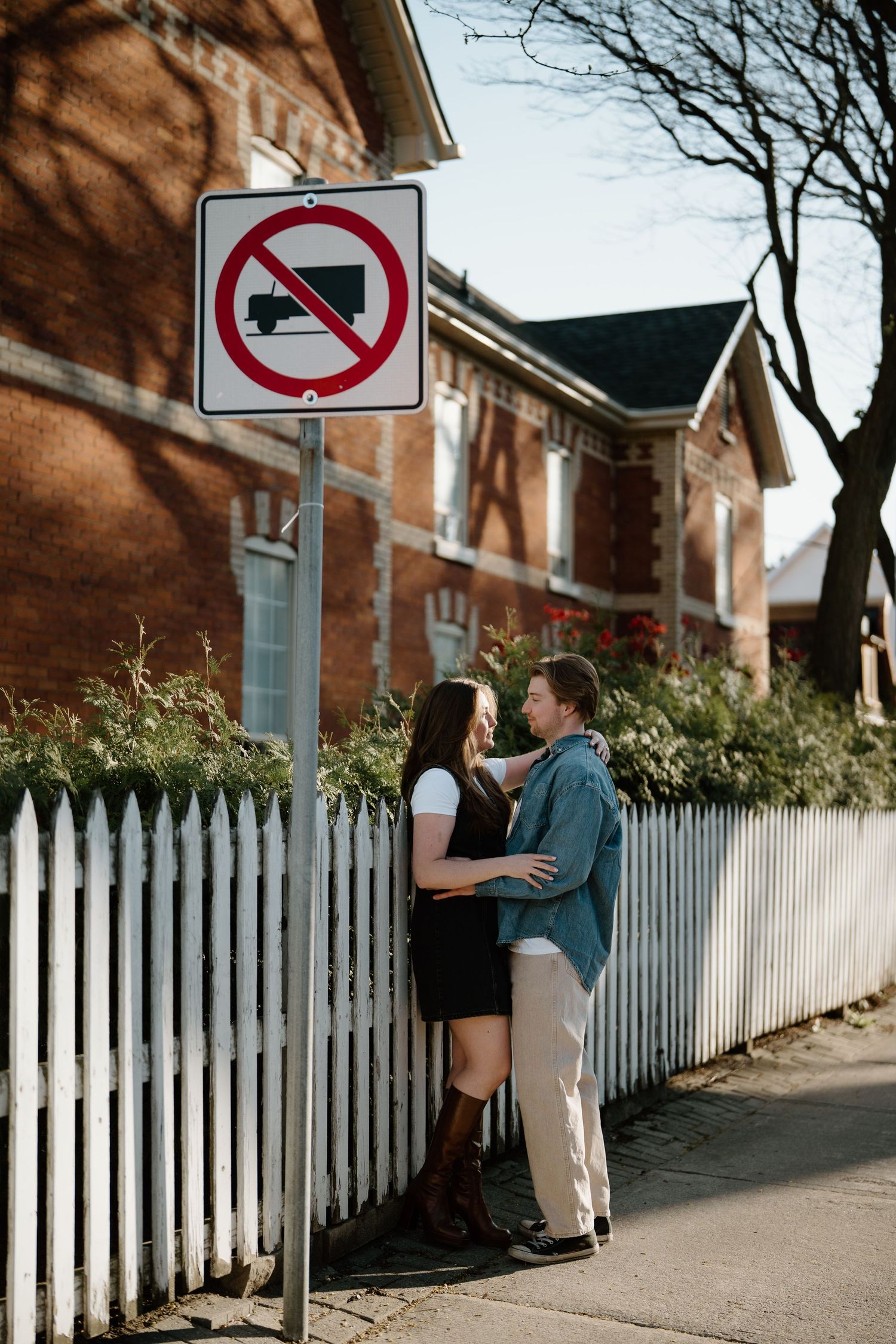 Two people share an intimate moment beside a white picket fence and brick building with a no trucks traffic sign above.