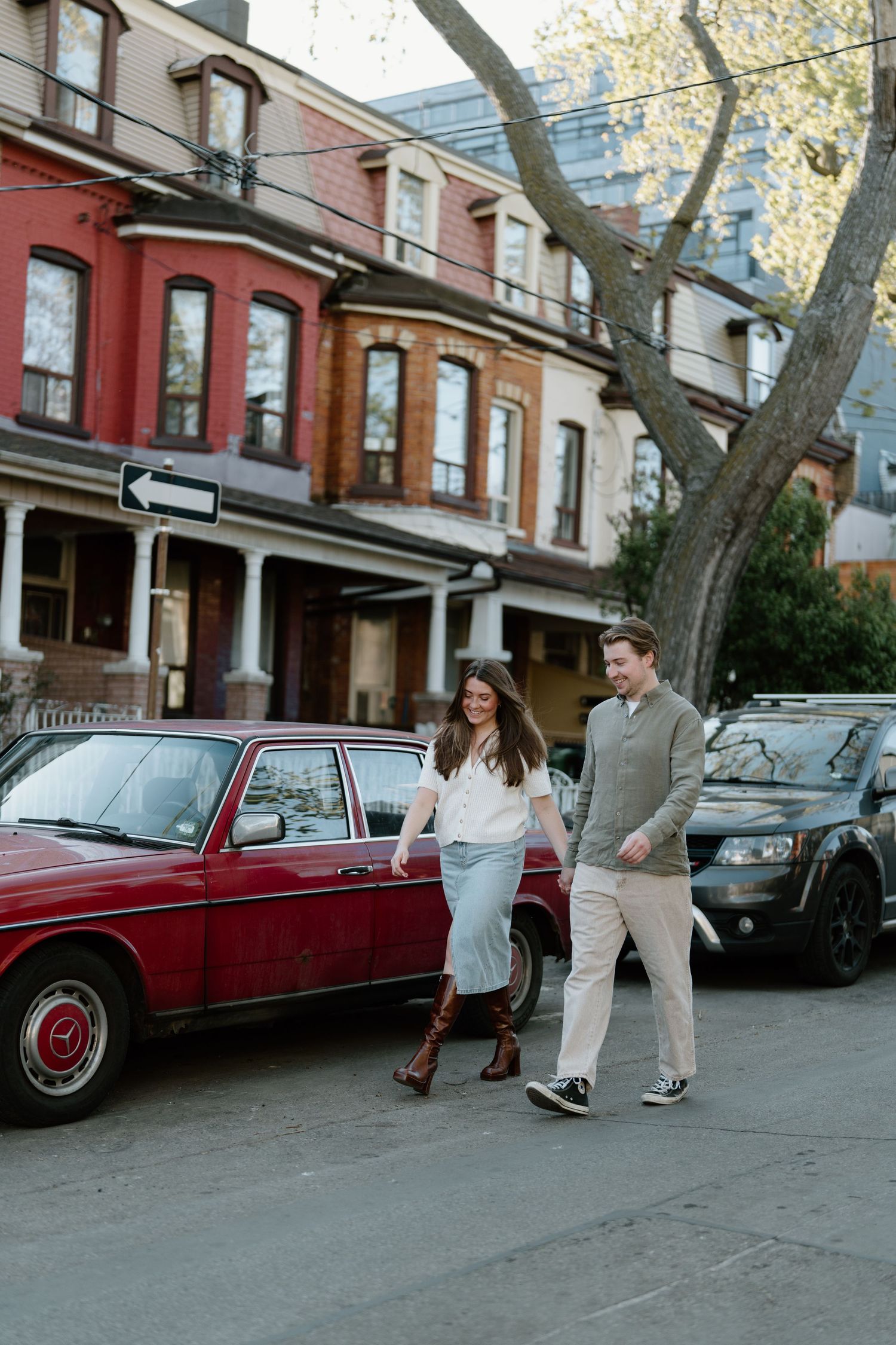 A couple walks down a historic street lined with Victorian row houses and parked cars including a vintage red sedan.