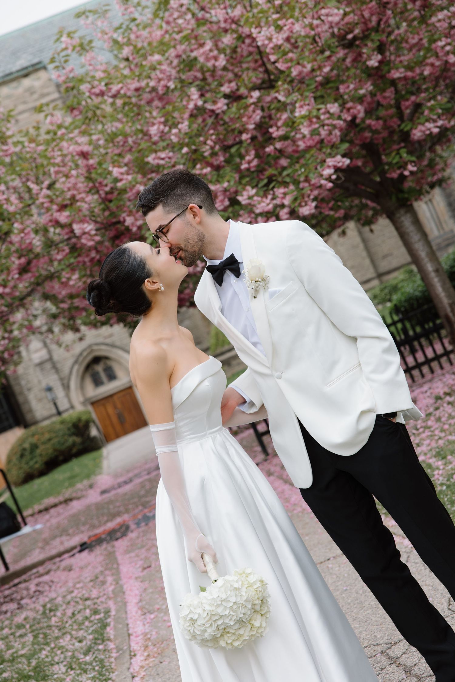 A couple shares a romantic kiss under blooming cherry blossoms while standing in front of a historic stone building.