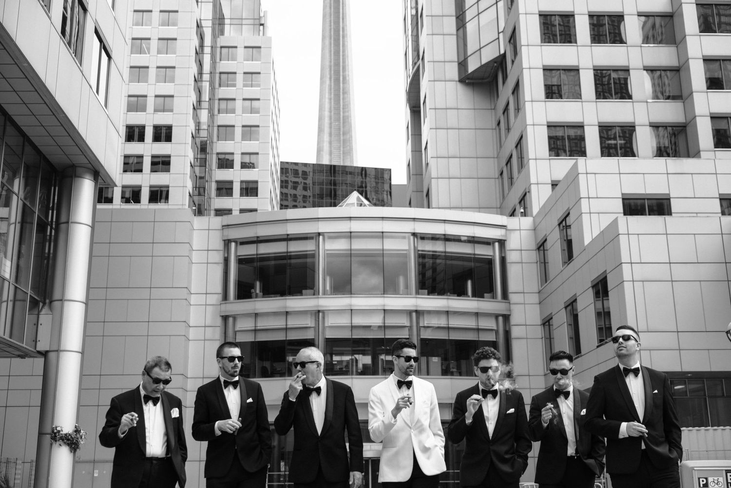 Stylish groomsmen in suits and sunglasses pose together against modern urban architecture backdrop in black and white.