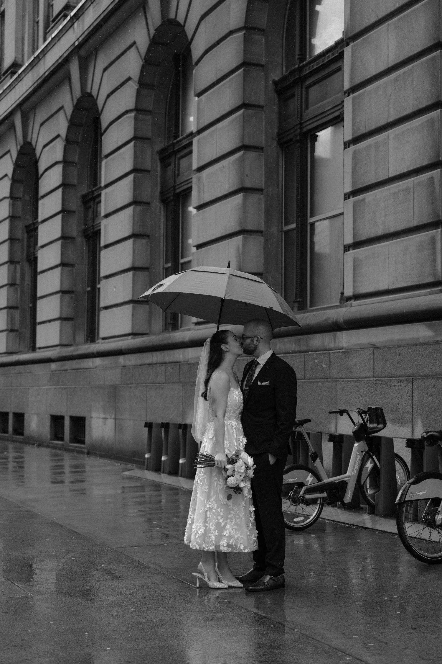 A couple shares an intimate moment under an umbrella on a rainy day outside a historic building in black and white.
