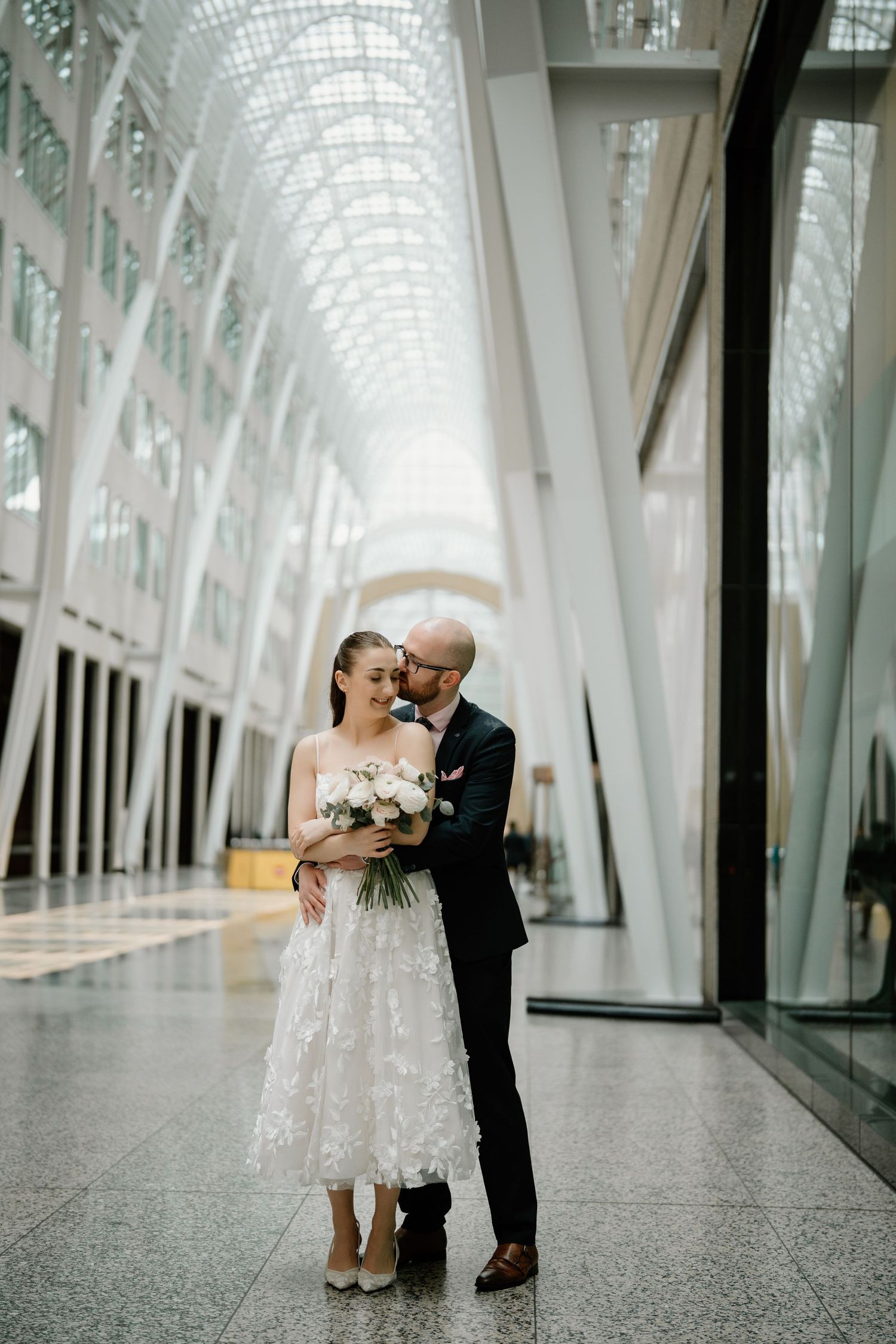 A romantic wedding couple embraces in a bright modern atrium with soaring glass ceilings and white architectural details.