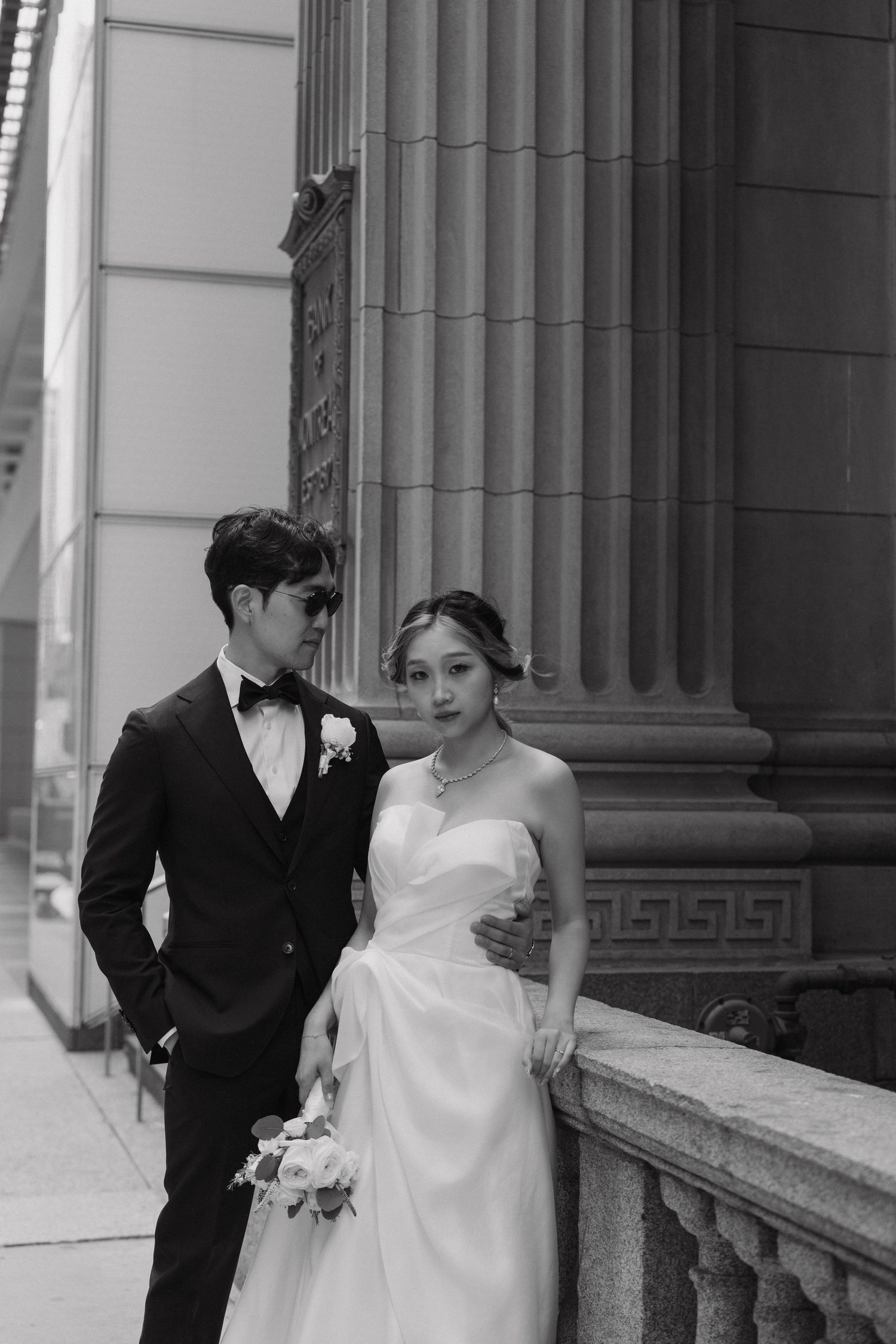 A black and white wedding photo of a couple posing along elegant architectural columns.