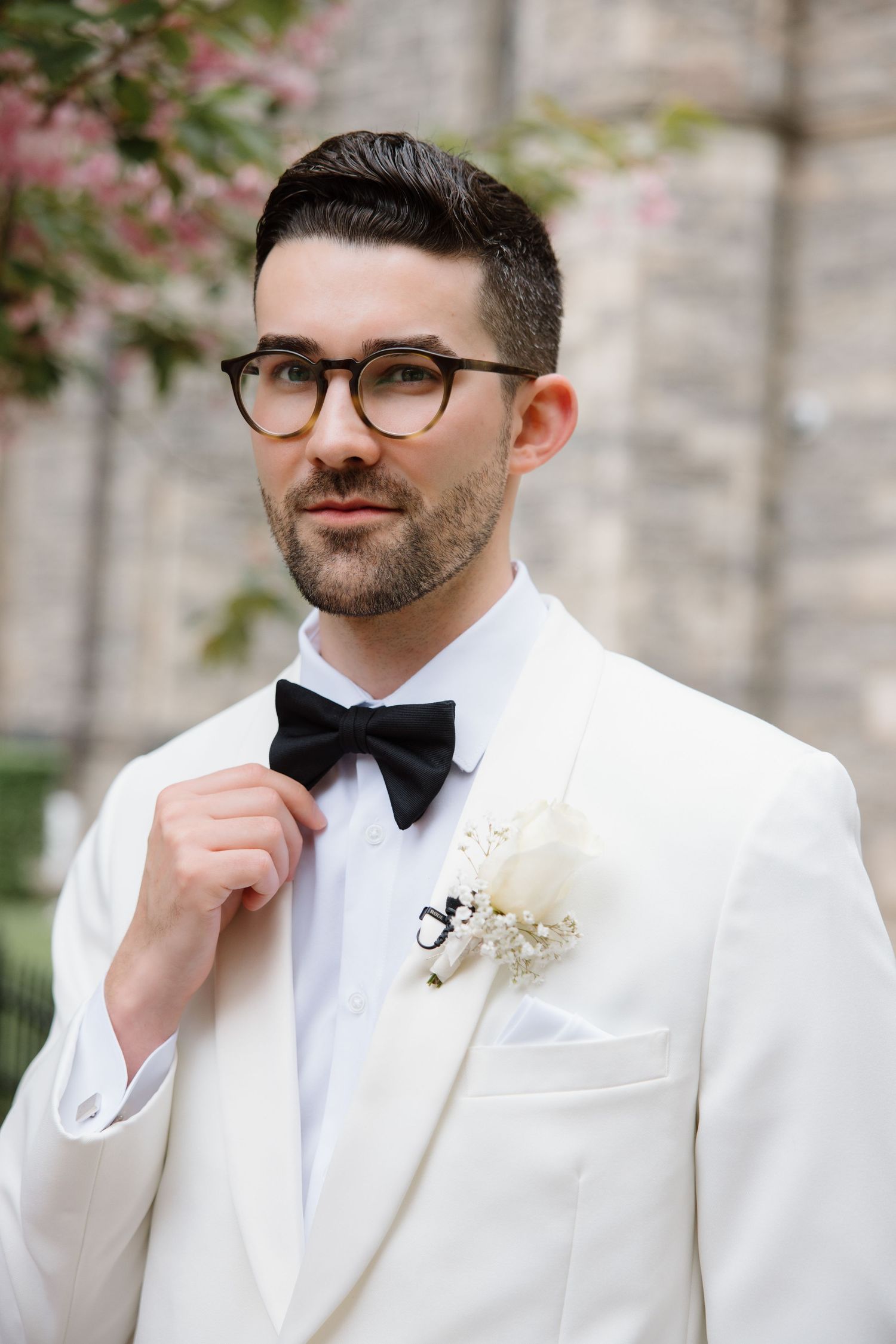 A groom in a white tuxedo jacket and black bow tie adjusts his tie while wearing glasses and a boutonniere.