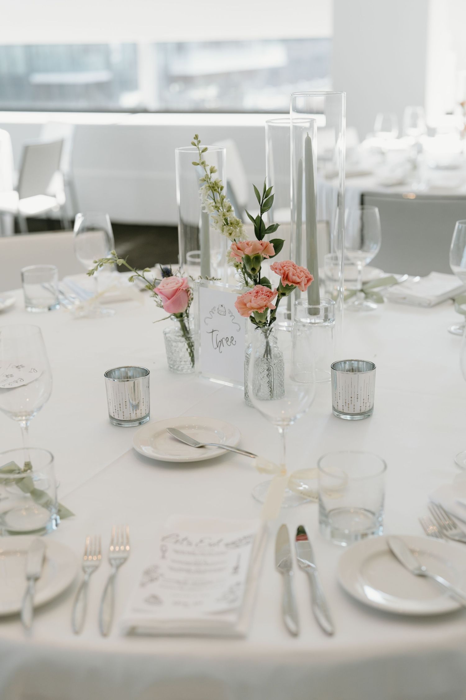 Elegant wedding table setting with white linens, pink flowers in small vases, and mercury glass candle holders.