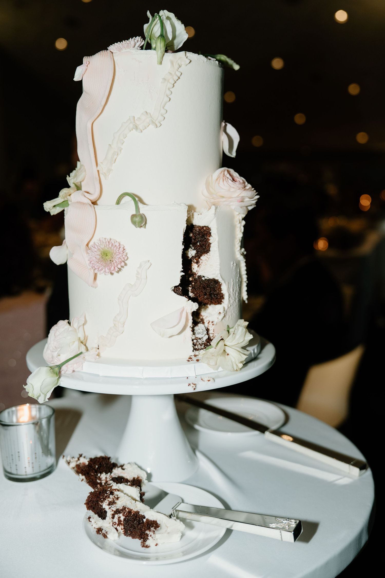 Elegant white wedding cake with floral decorations and a slice missing revealing chocolate interior on white pedestal stand.