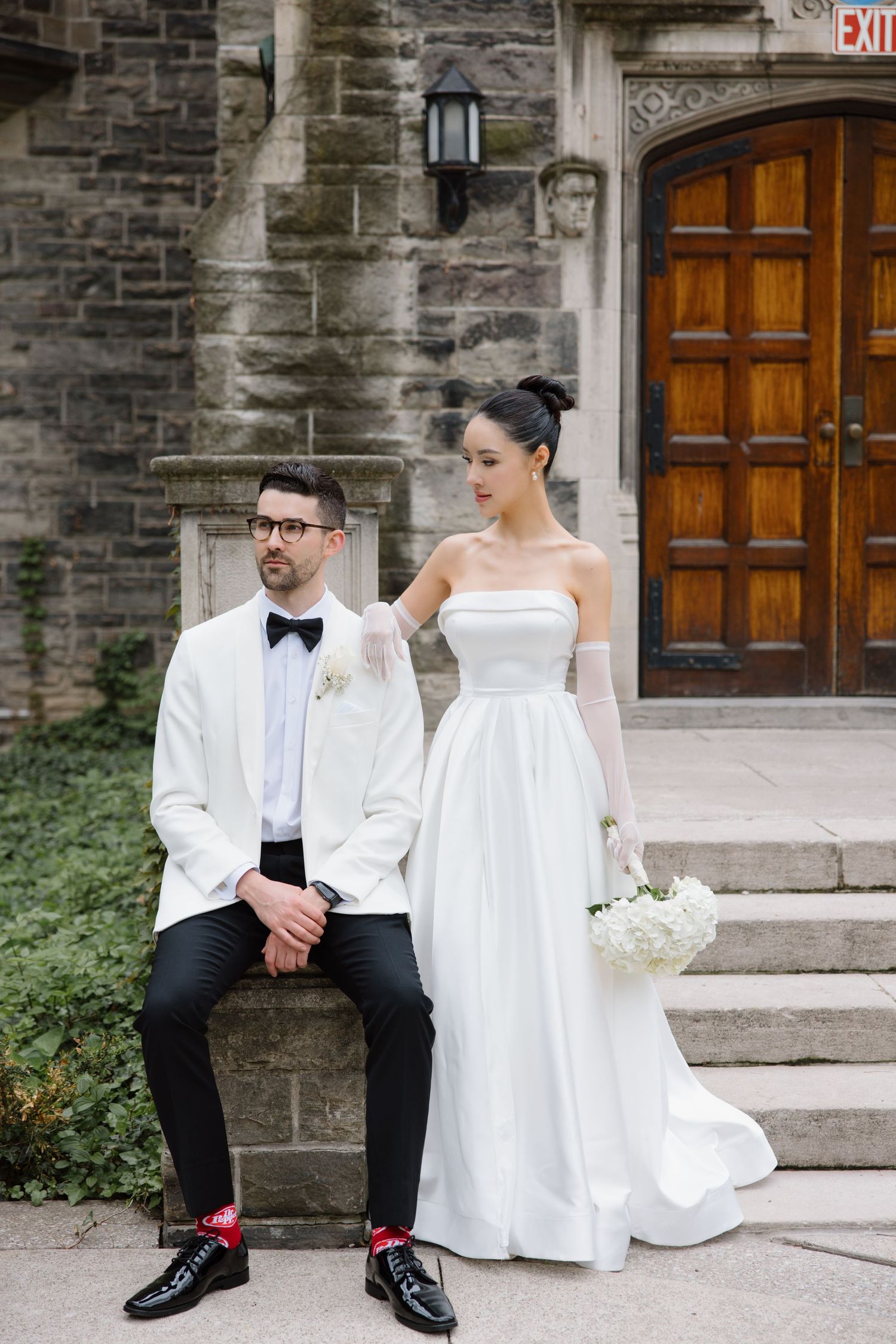 Stylish couple in wedding attire pose on stone steps with white dress and tuxedo jacket against church entrance.