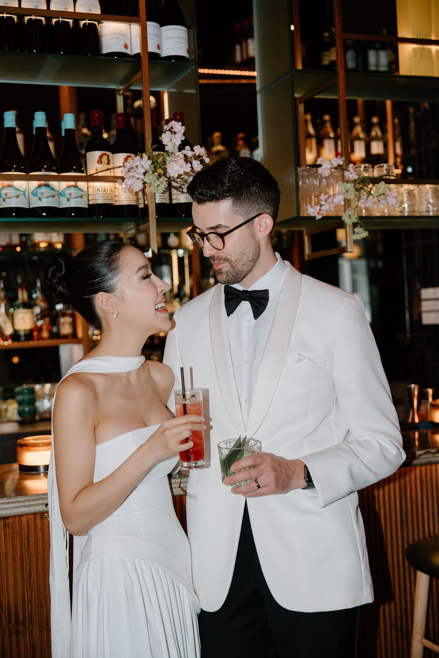 A couple in white formal attire share drinks together at an elegant bar with shelves of bottles in the background.