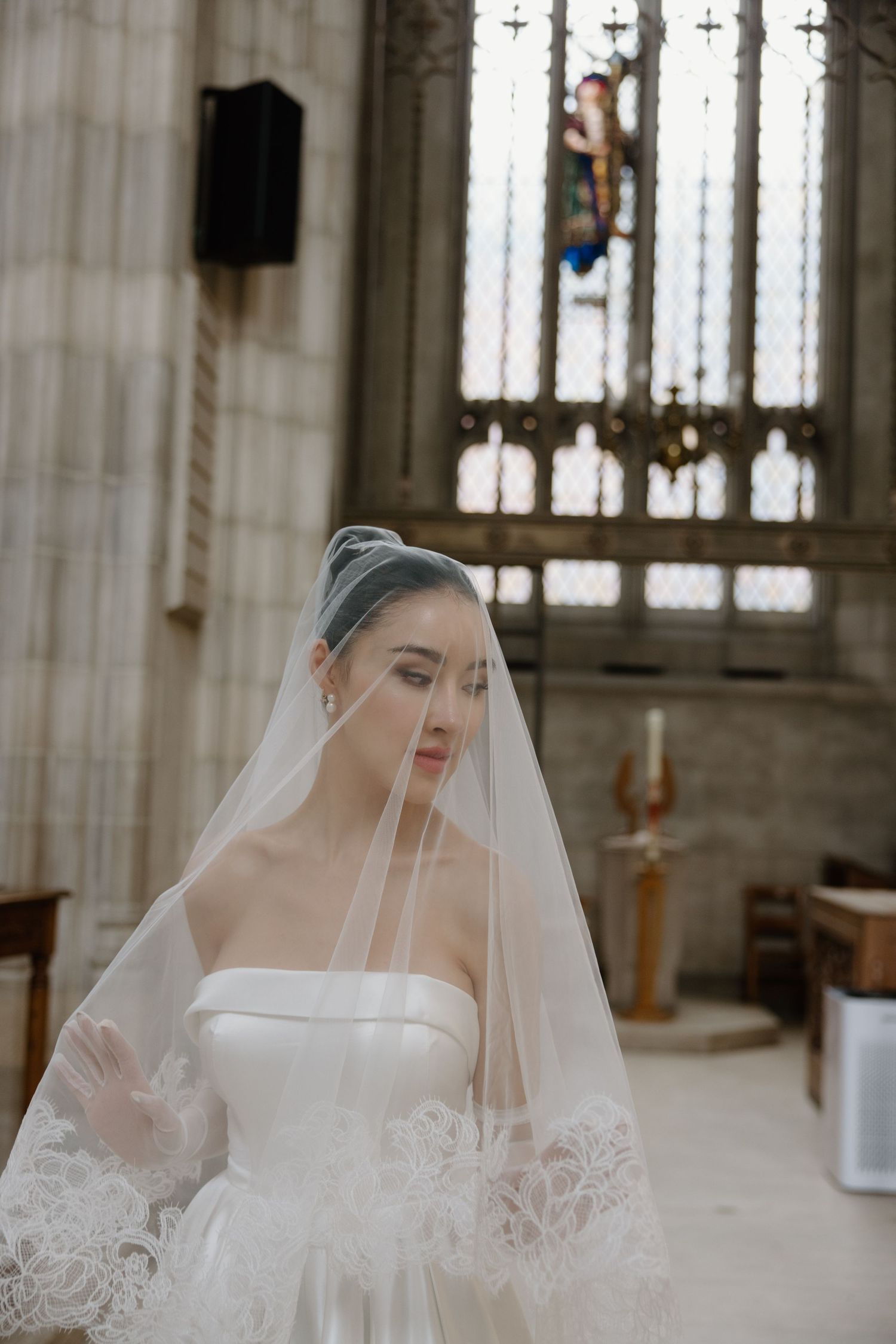 Bride wearing delicate white lace veil poses thoughtfully in ornate cathedral with stained glass windows and columns.