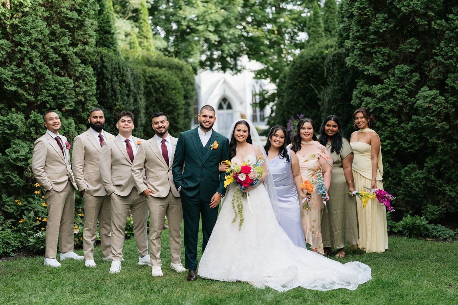 Wedding party in tan suits and yellow dresses poses together in a lush garden setting for formal portraits.