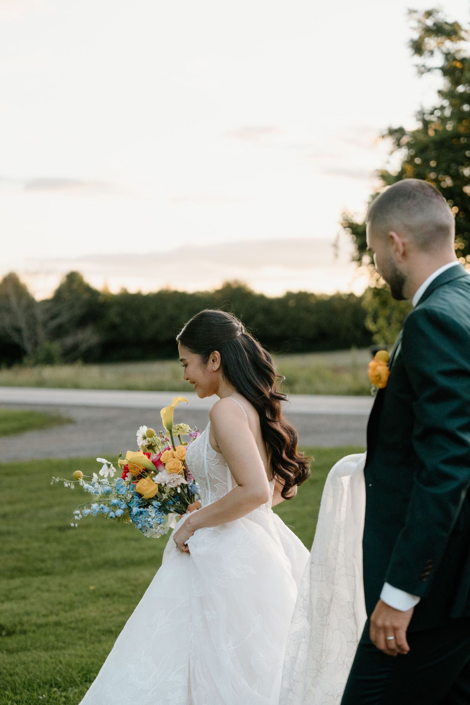 A couple in wedding attire walks hand in hand across a grassy field during sunset.